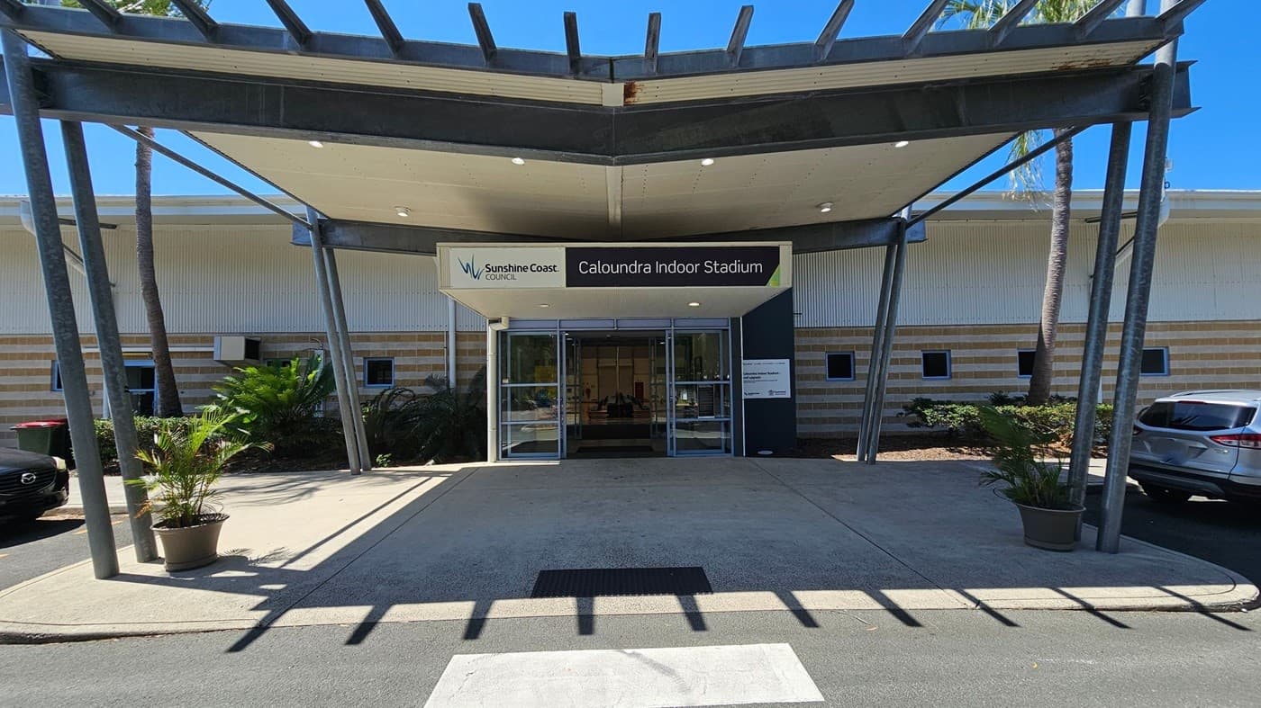 Entrance to Caloundra Indoor Stadium with a sign above the glass doors. The building has a striped exterior with small square windows. A metal awning supported by angled beams extends over the entrance. Potted plants are on either side of the walkway. Cars are parked on the right, and there are palm trees in the background. The ground is paved, and there is a pedestrian crossing in the foreground.