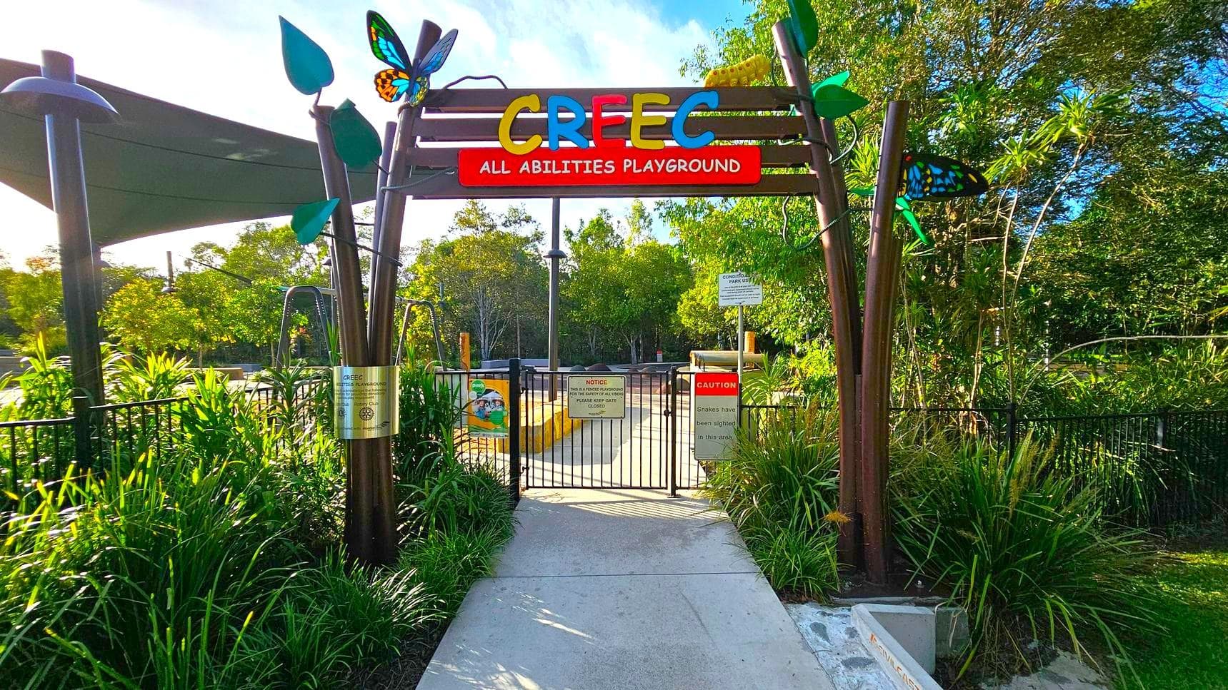 Entrance to a playground with a colourful sign reading "CREEC All Abilities Playground" at the top. The gate is black metal with various signs, including a caution about snakes. Decorative elements like butterflies and leaves adorn the entrance. Lush greenery surrounds the area, with trees and plants on both sides. A shaded area with a canopy is visible on the left. The path leading into the playground is concrete.