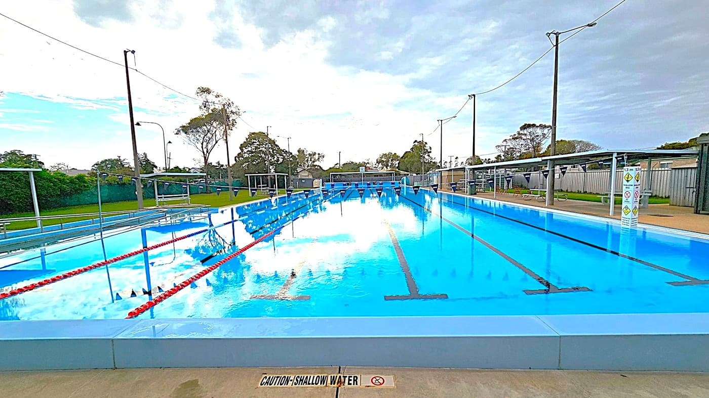 Outdoor swimming pool area with a covered seating section on the left, featuring several plastic chairs. In the centre, a sign with safety instructions is visible. The pool is on the right, with clear water and lane dividers. Trees and some buildings are in the background. The ground is a mix of green and beige concrete, and the pool has metal handrails. The sky is overcast with patches of blue.