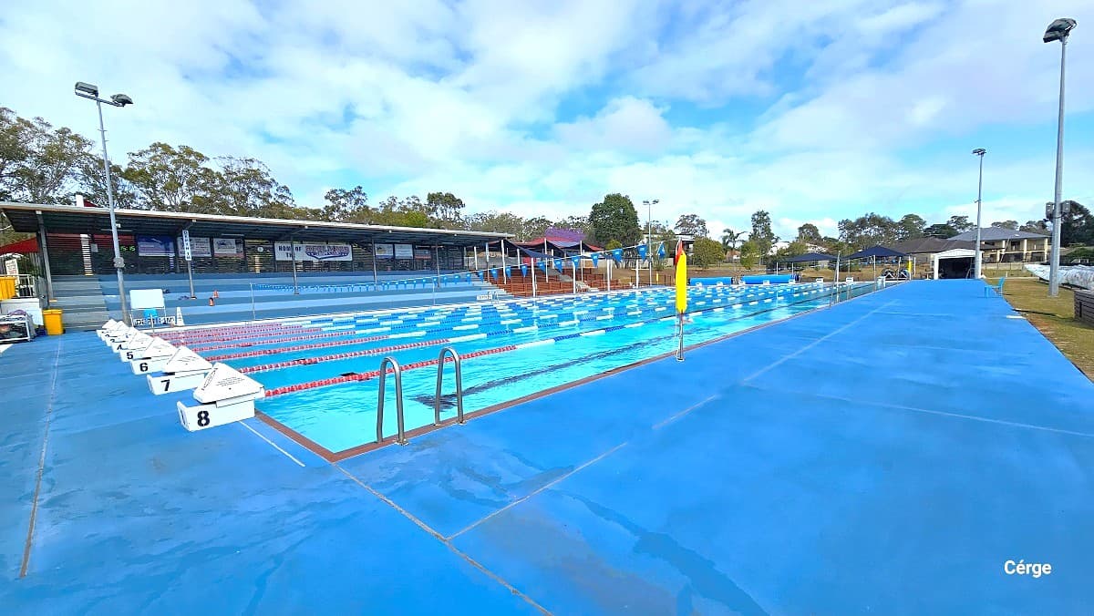Cleveland Aquatic Centre has a 50m outdoor pool with a wide pool deck. On the right side is brown-tiered seating with a black sunshade and blue-tiered seating with a solid roof. Behind it are low-set brick cream-coloured buildings with brown tiled roofing having solar panels. Pool lanes are marked by white diving blocks marked from 1 to 8 and are separated by red, blue, and white floating rope lanes.