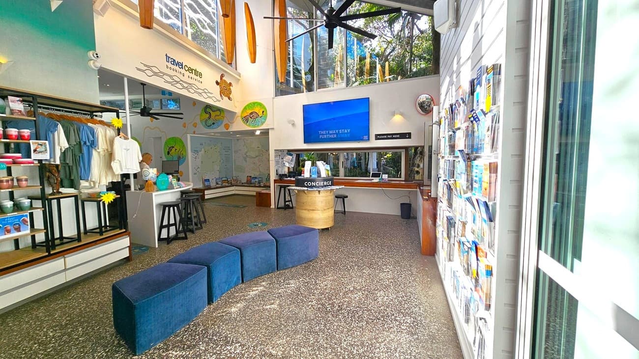 A visitor information centre with a modern design. On the left, there is a display area with brochures, a rack of T-shirts, and a sign for "Sunny Jeeps". In the centre, a glass door is flanked by posters about community projects and plastic-free initiatives. On the right, a service window with brochures and a digital screen. The ceiling has wooden panels, and the floor is tiled in various shades of green and brown.