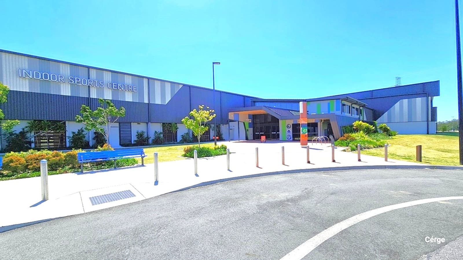 The South Pine Indoor Sports Centre, situated on South Pine Road in Brendale, is a spacious building with grey corrugated iron and vibrant accents. Exterior signs indicate 'Rugby Union,' 'Touch Football,' and 'Indoor Sports Centre.' Bike racks and seating are near the entrance. The facility is surrounded by open spaces and gardens.