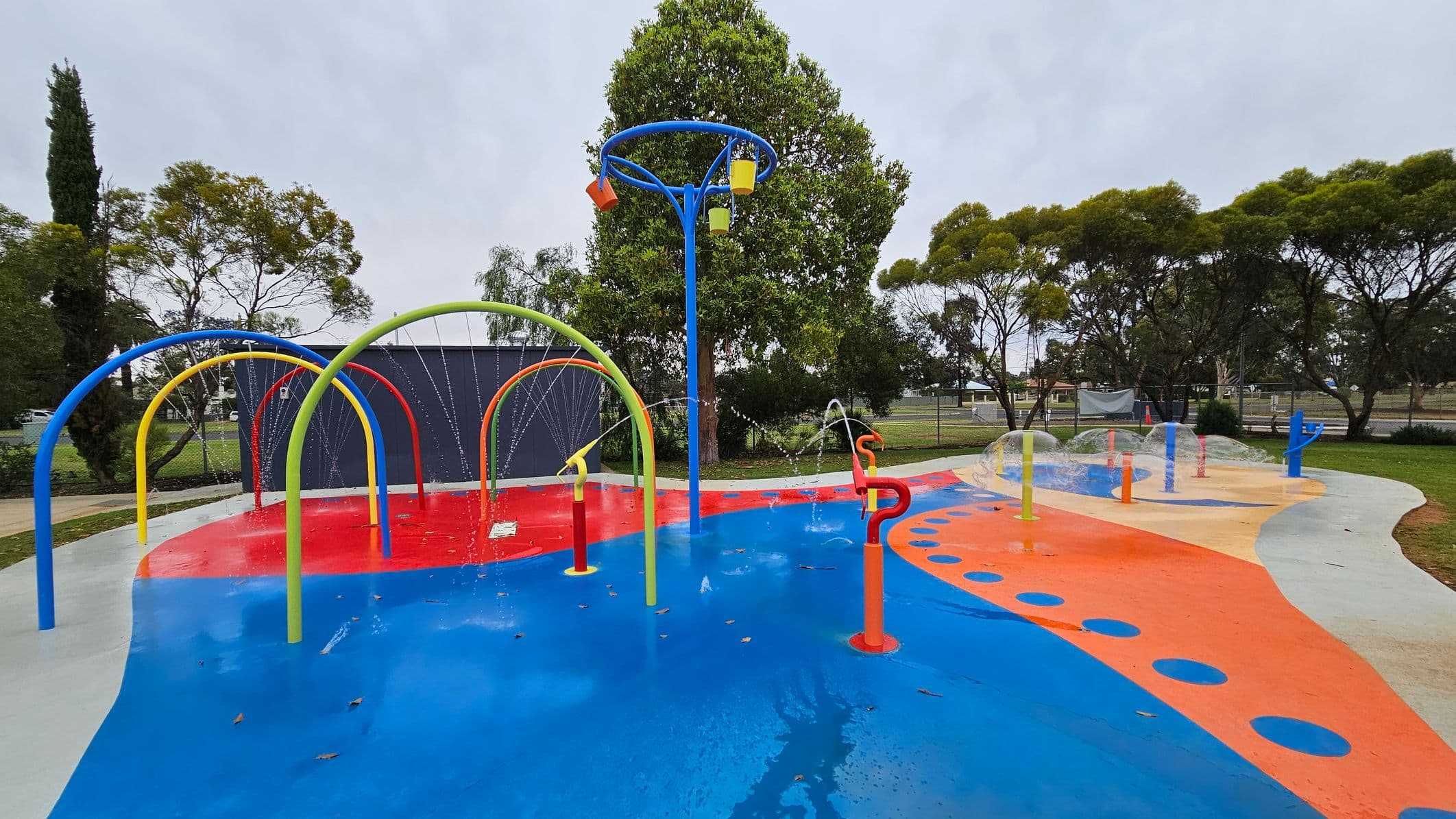 A colourful splash pad with various water features. On the left, there are blue, yellow, and red arches spraying water. In the centre, a tall blue structure with buckets that tip water. On the right, orange and blue poles with water jets. The ground is painted in bright blue, red, and orange patterns. Trees and a fence are in the background, with a large tree in the centre. The area is surrounded by grass.