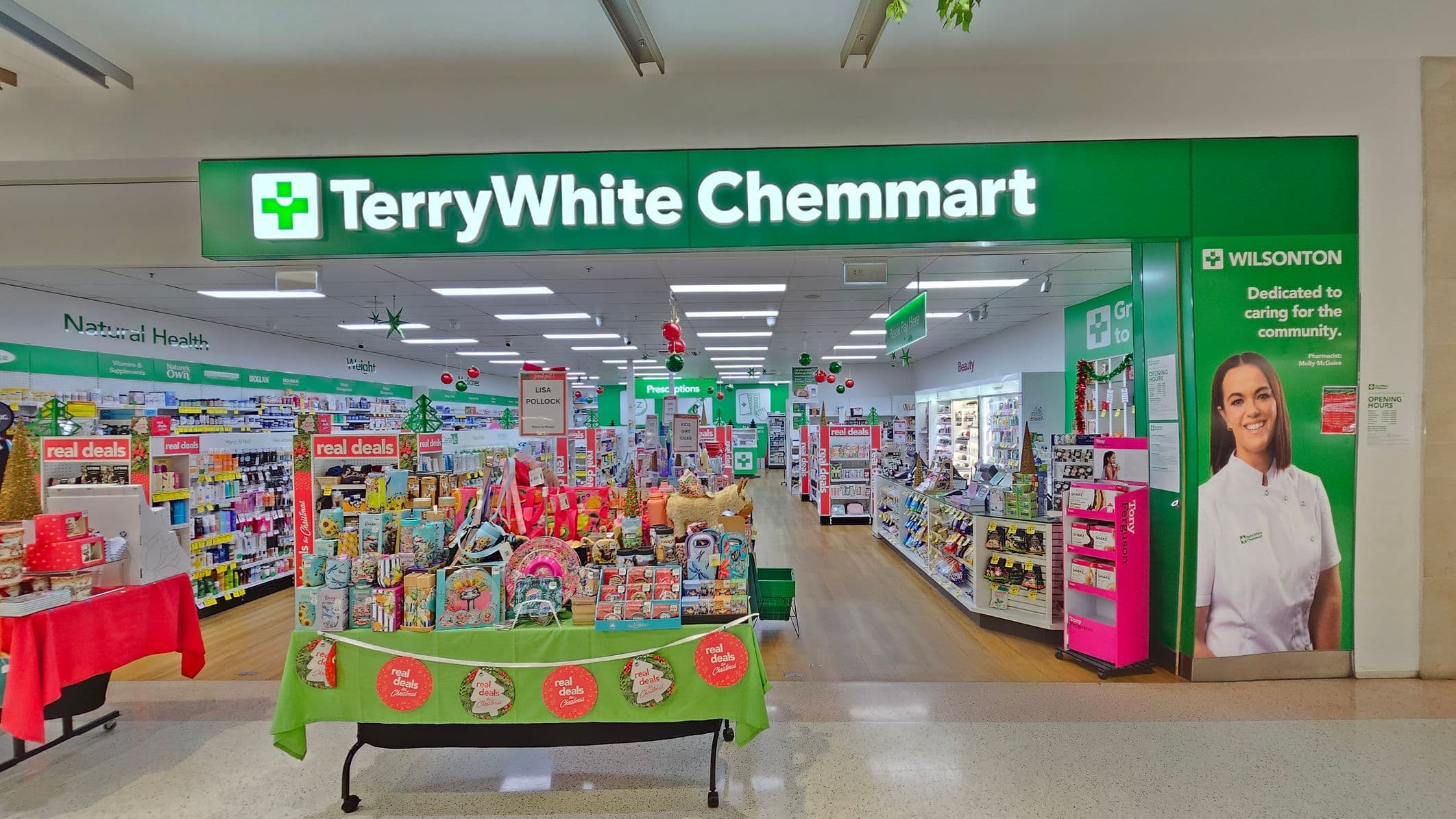 Entrance to a pharmacy with a green sign reading "TerryWhite Chemmart" above. Inside, shelves are stocked with various products, including health and beauty items. A display table in the centre features promotional items with a green and red theme. On the right, a large poster of a smiling person in a white uniform is visible. The floor is a mix of wood and light-coloured tiles, and the ceiling has fluorescent lighting.