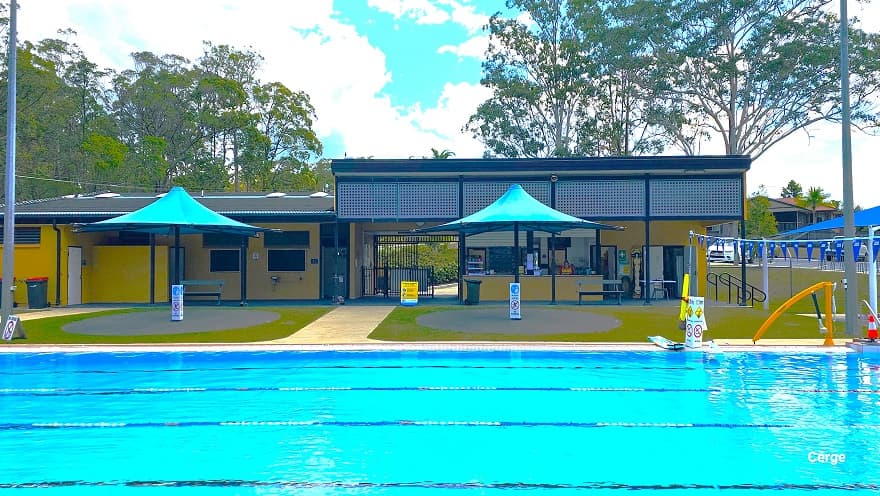 6-lane outdoor lap pool at Woodford. At the starting end are white diving blocks numbered from 1 to 6 using blue paint. Behind the pool deck of the starting section is a blue dome shade with grey benches and picnic-style tables. Further to the left are low-set orange buildings with glass windows and four blue sunshades. The other end of the pool is a dark grey corrugated iron shed for storage. Tall gum trees surround the property