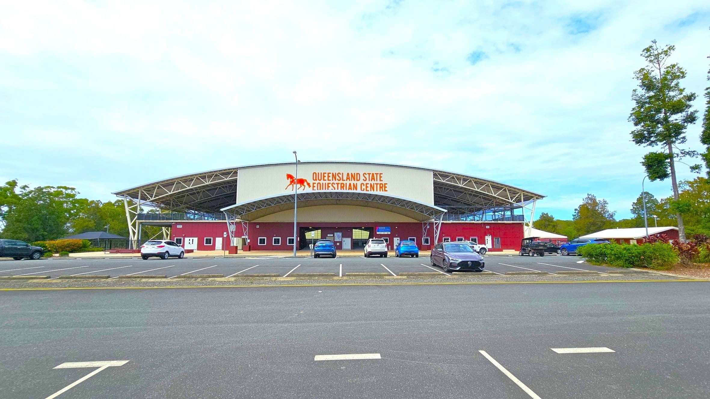A large stadium structure with a metal roof and white support beams on the left. The building has a maroon facade with several white doors and windows. In the foreground, there are red benches and rectangular planters on a paved area. A black metal fence is visible near the entrance. On the right, a road with a parked vehicle and portable buildings can be seen. Trees are in the background.