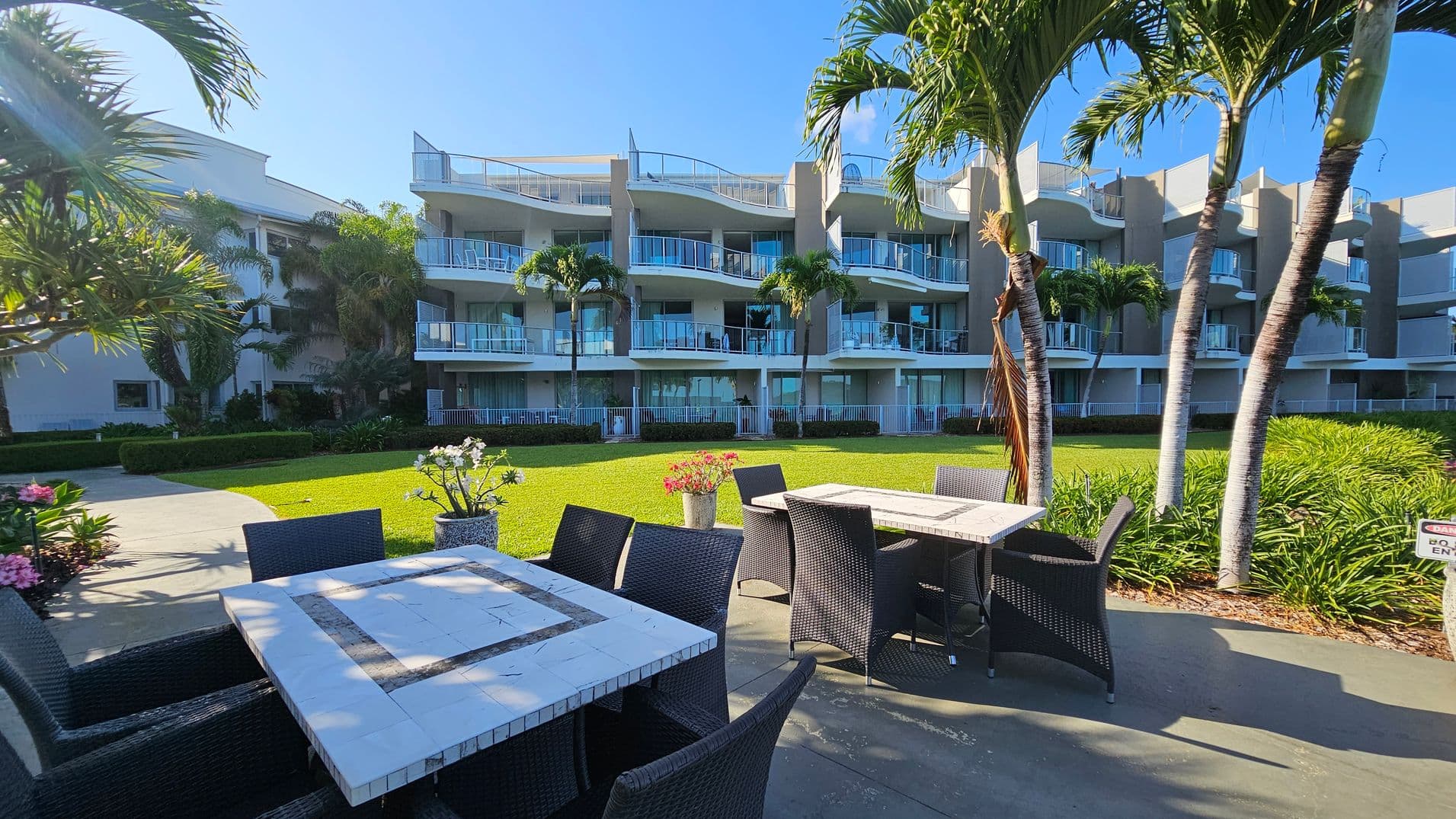 Outdoor seating area with two square tables and wicker chairs on a paved surface in the foreground. Potted plants with flowers are placed near the tables. In the background, a modern multi-storey building with balconies and glass railings is visible. Palm trees and well-maintained grass surround the area, adding a tropical feel.