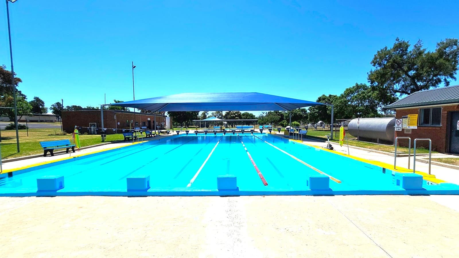 A rectangular swimming pool with clear blue water is in the centre, featuring lane dividers. A large blue shade structure covers part of the pool. Surrounding the pool are concrete walkways and grassy areas. On the left, there are blue benches and a small building. On the right, a cylindrical metal tank and another building are visible. Trees and open space are in the background.