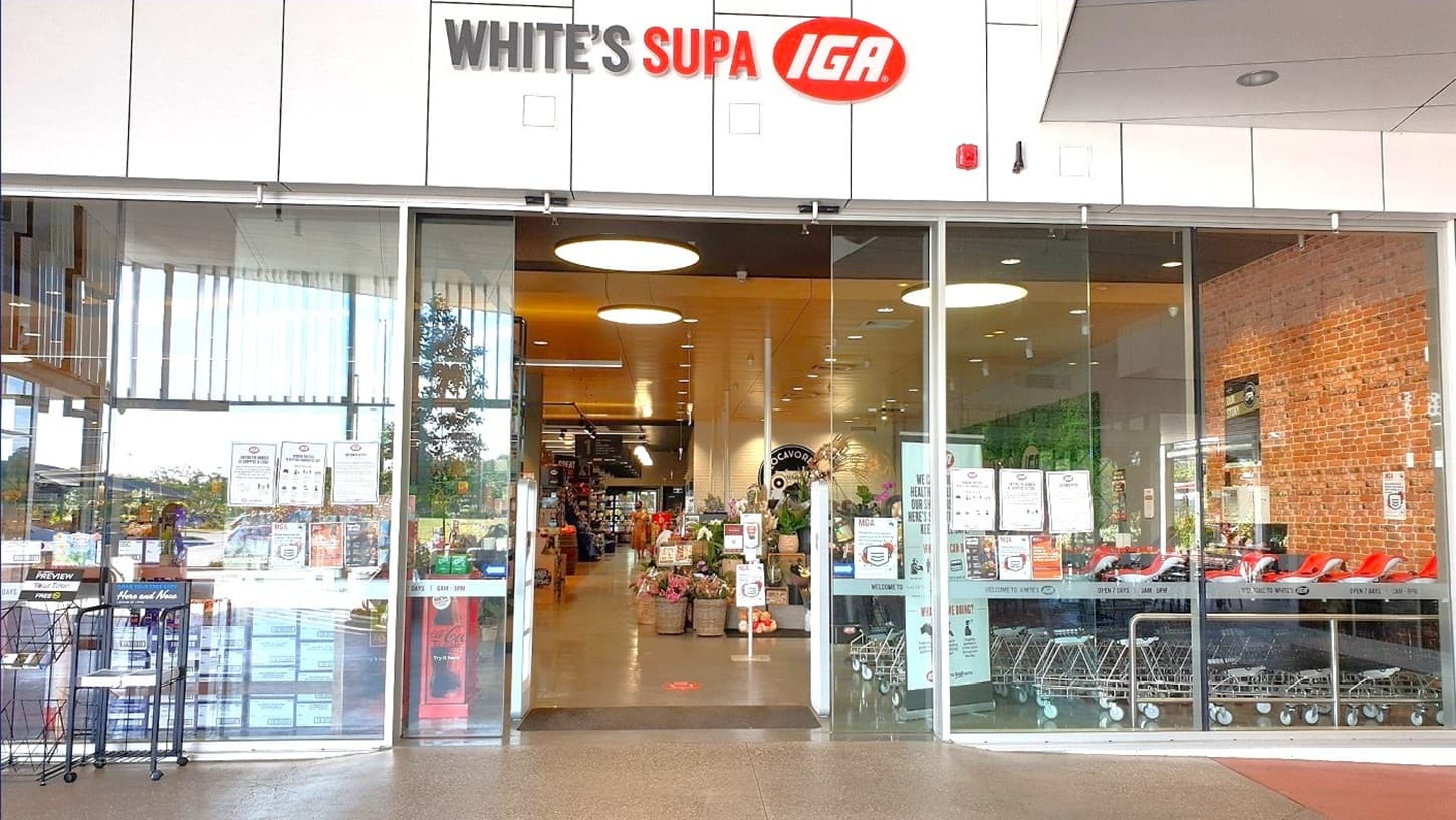 The image shows open glass doors at White’s IGA Baringa, which is regulated by a motion detector. The building has white walls with black lines that make it look like it is made of tiles. The wall on top of the glass doors is the building name with its brand colors which is White’s Supa IGA. On the left side are black magazine racks. Visible through the glass doors are the trolleys, the display stand of flowers, and the aisle leading to other sections inside the store.