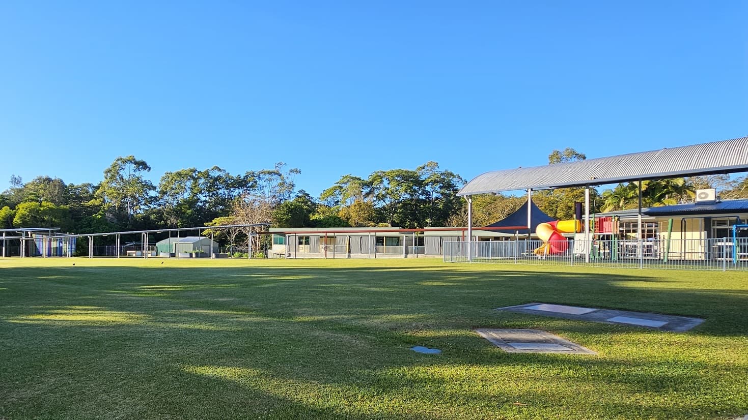 On the left, there are tall trees and bushes bordering a large grassy field. In the middle, there is an expansive lawn with a few scattered concrete utility covers. On the right, there are several buildings with covered walkways and a playground area featuring a red and yellow play structure under a black shade sail. The floor is primarily grass, and the ceiling of the covered walkways is metal with exposed beams. The background includes more trees and additional buildings.