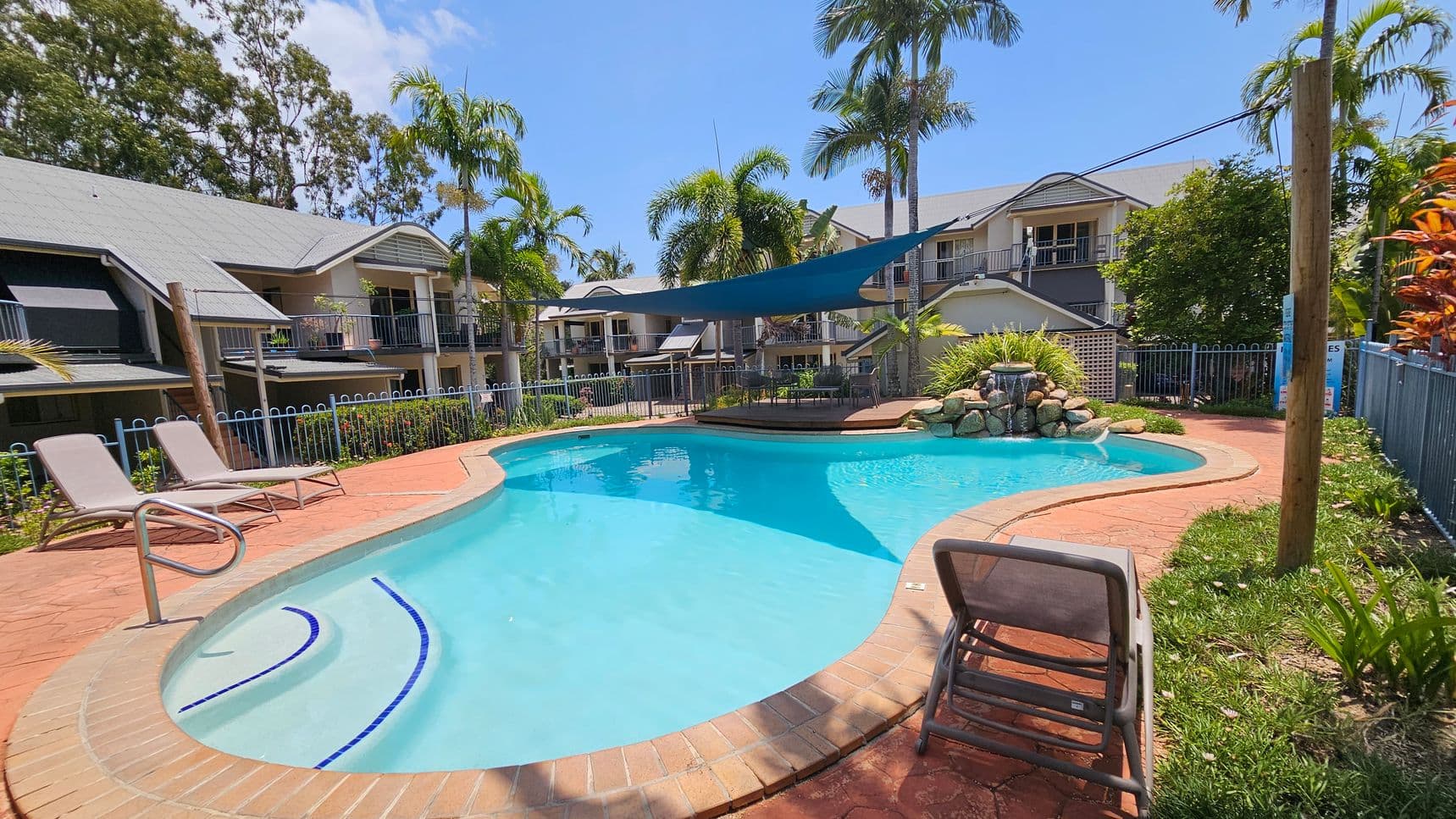 A swimming pool with clear blue water is surrounded by a red brick patio. On the left, two sun loungers are positioned near the pool's edge. In the background, there are several two-storey buildings with grey roofs and balconies. Palm trees and other greenery are scattered around the area. A blue shade sail is stretched over part of the pool, and a small waterfall feature is visible at the far end. A fence encloses the pool area.