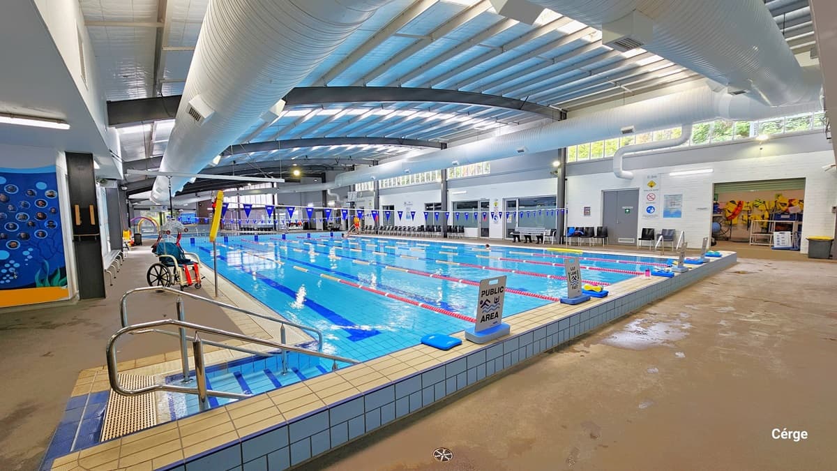 25-metre Heated Indoor Pool at Monbulk Aquatic Centre. Near the white tile pool at the starting line, to the left, are stairs with metal handrails. The pool deck is concrete in color. The facility has white walls, large white ventilation ducts, and grey metal beams in the ceiling. On the upper right wall are small glass windows that allow natural light to filter in. Metal benches and black plastic chairs are available around the pool deck. There is an aquatic chair on the left pool deck.