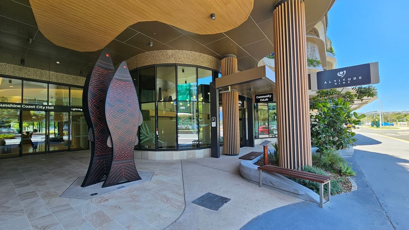 Entrance to a modern building with large glass windows and a curved design. On the left, there's a sculpture of two stylised fish with geometric patterns. The floor is paved with light-coloured tiles. On the right, a wooden bench and a sign for "Altitude Nine" are visible. A potted plant and a small garden area add greenery. The ceiling has a wooden finish with recessed lighting.