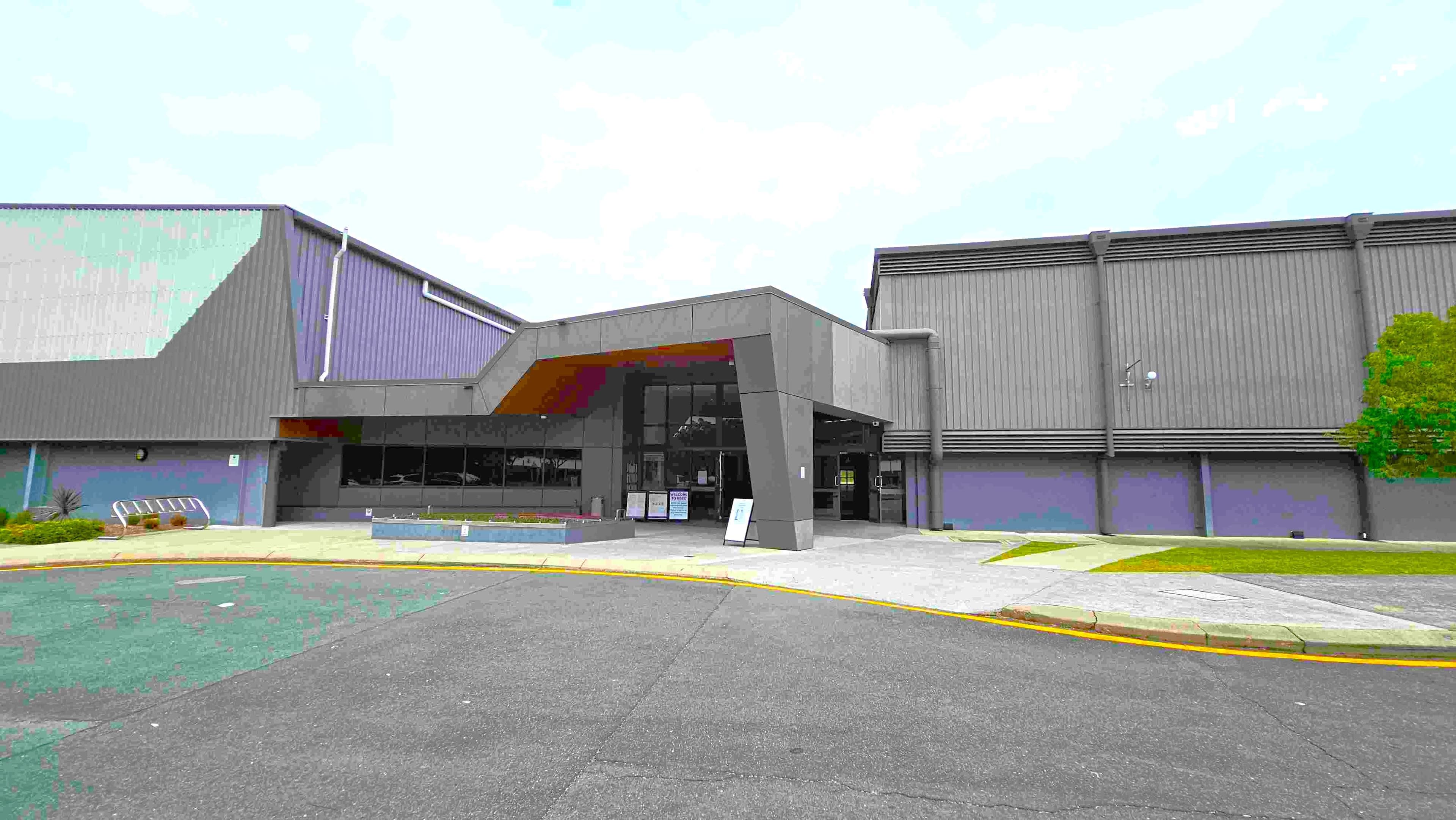 On the left, a large grey building features a sloped roof with vertical panels and a smooth facade. The entrance is highlighted by a modern overhang and large glass doors. A small landscaped area with low plants is visible in front of the entrance. To the right, the building continues with a similar grey exterior, and a small tree adds greenery. The ground is asphalt, with a curved pathway leading towards the entrance.