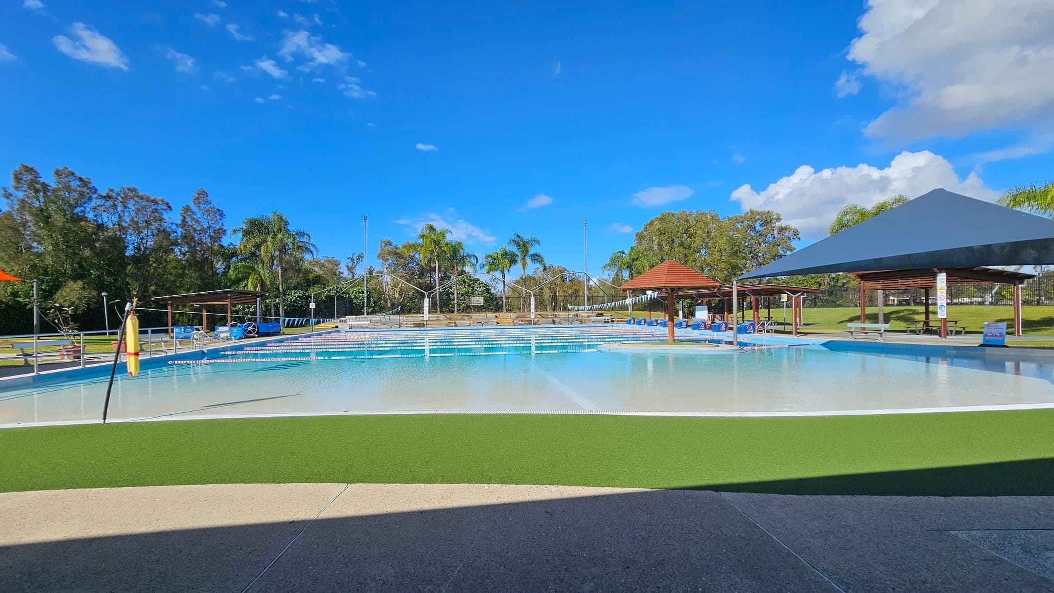 A large outdoor swimming pool with clear water is surrounded by a concrete deck and artificial grass. On the right, a blue shade structure covers a seating area with picnic tables. In the centre, a small wooden gazebo provides additional shade. Palm trees and other greenery line the background, with a clear blue sky above. The pool area is equipped with safety ropes and lifeguard equipment.