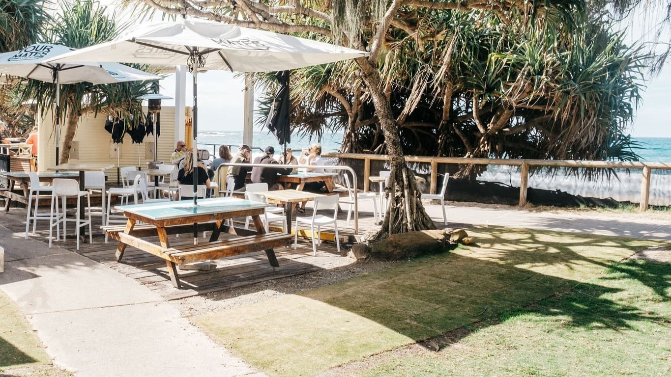 On the left, there are white umbrellas over tables and chairs, with people sitting and a bar counter with stools nearby. In the middle, there is a large tree with a thick trunk and sprawling branches, surrounded by more tables and chairs, including picnic tables. On the right, there is a wooden fence with dense foliage and the ocean visible beyond it. The floor is a mix of concrete and grass, and the ocean is visible on the right.