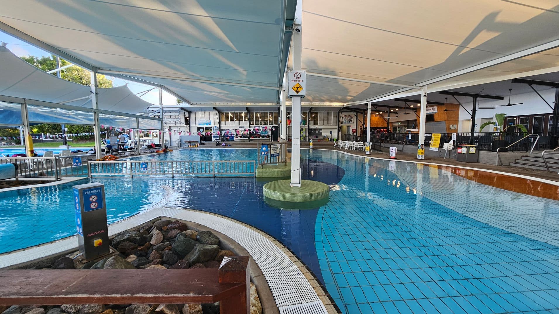 Indoor swimming pool area with a tiled floor and a curved pool. On the left, there are lounge chairs and a small fence separating the pool sections. A sign with spa rules is visible near a rocky area. The ceiling is covered with large white fabric canopies. On the right, there is a seating area with tables and chairs, and a café counter in the background. The area is well-lit with overhead lights, and there are signs and safety notices around the pool.