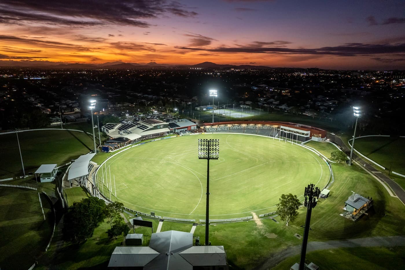 Aerial view of a sports stadium with a circular green field in the centre, surrounded by tall floodlights. On the left, there are pathways and parked cars, while on the right, buildings and additional parking spaces are visible. In the foreground, a large building with a red roof curves around the field. The background features a sprawling landscape with houses and open fields. The floor consists of paved walkways and grass patches.
