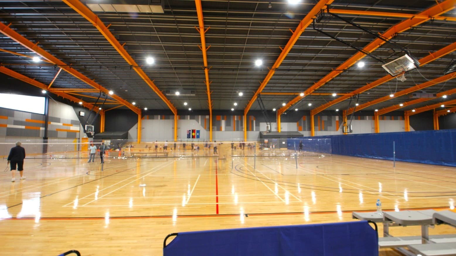 Inside the stadium of the Somerville Recreation Centre. The venue has a grey ceiling and bright yellow metal archways. It has polished light pine timber flooring with black, white, and red lines for different sports events. The lights coming from the fluorescents on the ceiling are seen to be reflected on the timber floor. The scorer's table is located in between the grey benches with orange lanes on the left side of the court, while on the opposite is 4-tier seating.