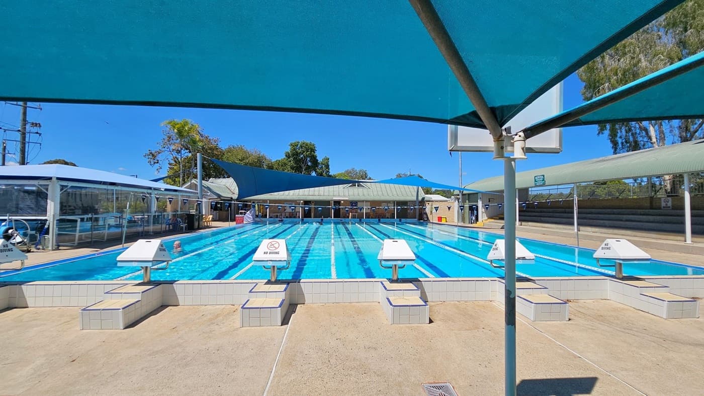 Outdoor swimming pool with multiple lanes, each marked by white and blue lane dividers. In the foreground, there are starting blocks with "No Diving" signs. The pool is covered by blue shade sails. On the left, there is a glass-enclosed area with a lifeguard chair. On the right, there are tiered seating stands. In the background, there are trees and a building with a green roof. The pool deck is concrete.
