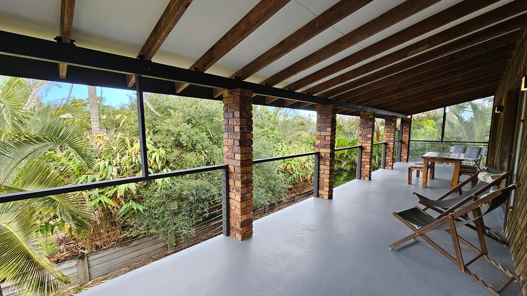On the left, there are tall green plants and trees visible through black-framed glass panels. In the middle, there are brick columns supporting the roof, with black metal railings and glass panels between them. On the right, there is a wooden table with chairs and a wooden bench. The floor is a smooth, light grey surface, and the ceiling has exposed wooden beams with white panels in between.