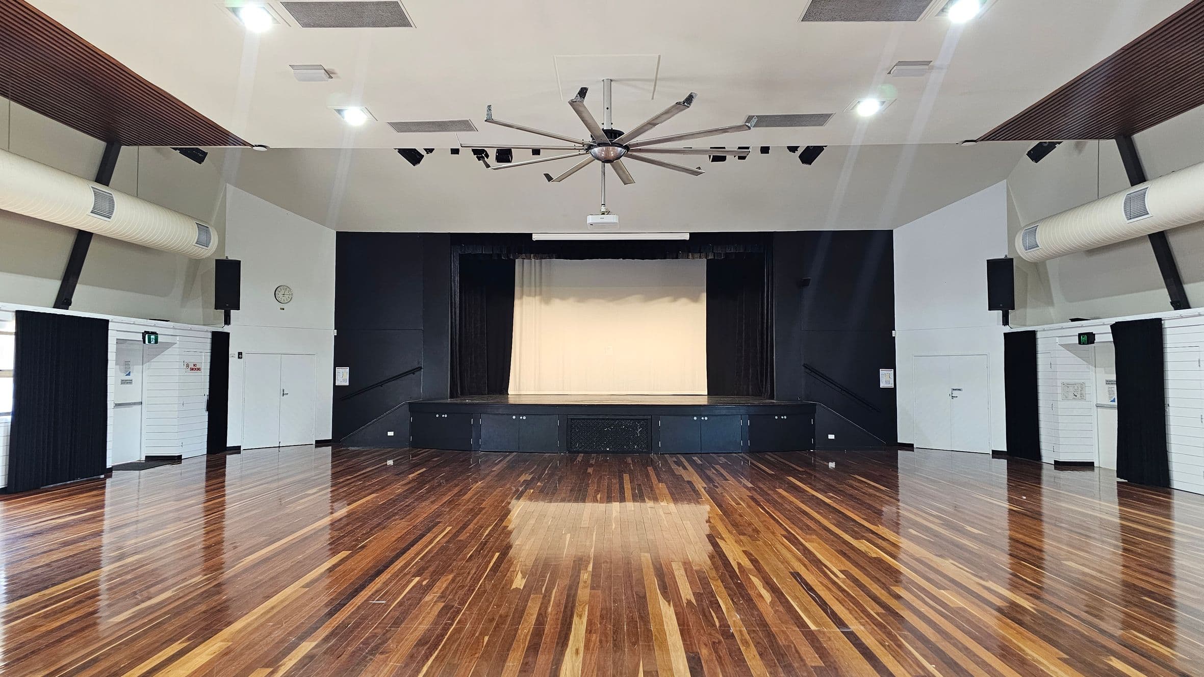 A spacious hall with a polished wooden floor and a stage at the far end. The stage has a light-coloured backdrop and is flanked by dark curtains. The ceiling features a large, central fan and several lights. On the left and right walls, there are air conditioning ducts and black curtains. A clock is visible on the left wall. The walls are light-coloured with some dark panels. There are doors on both sides of the room.