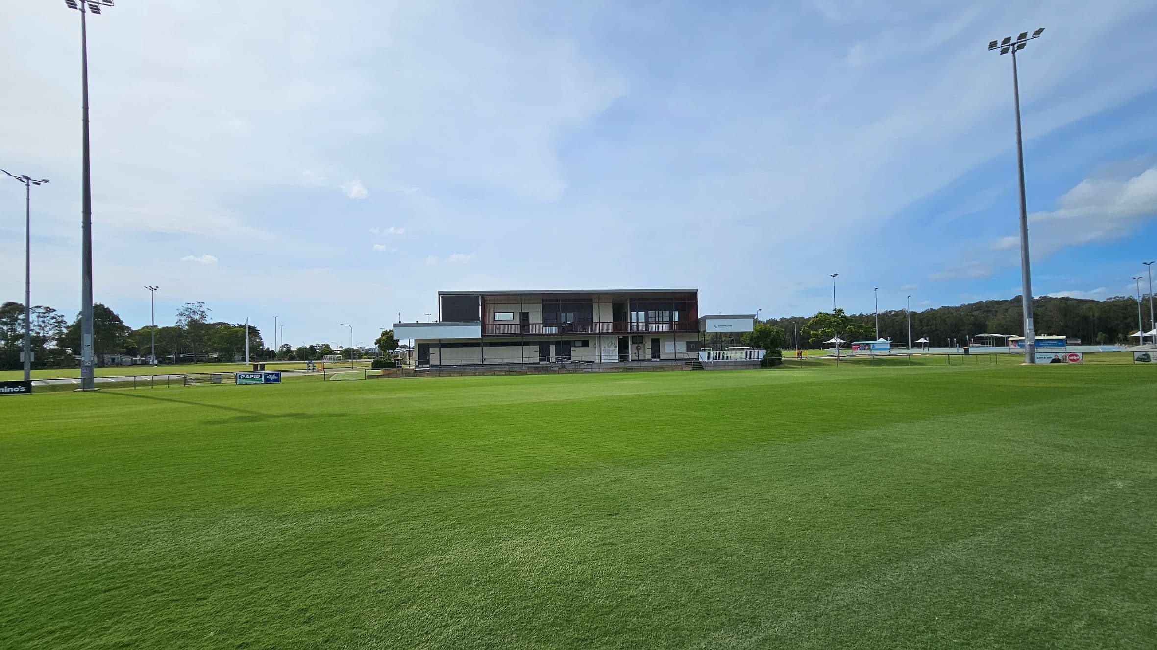 A sports field with well-maintained green grass. In the centre, there is a modern, two-storey building with large windows and a balcony. Tall floodlights are positioned on both the left and right sides of the field. In the background, there are trees and additional structures, possibly part of a sports complex. The area is open and spacious, with clear visibility of the surroundings.