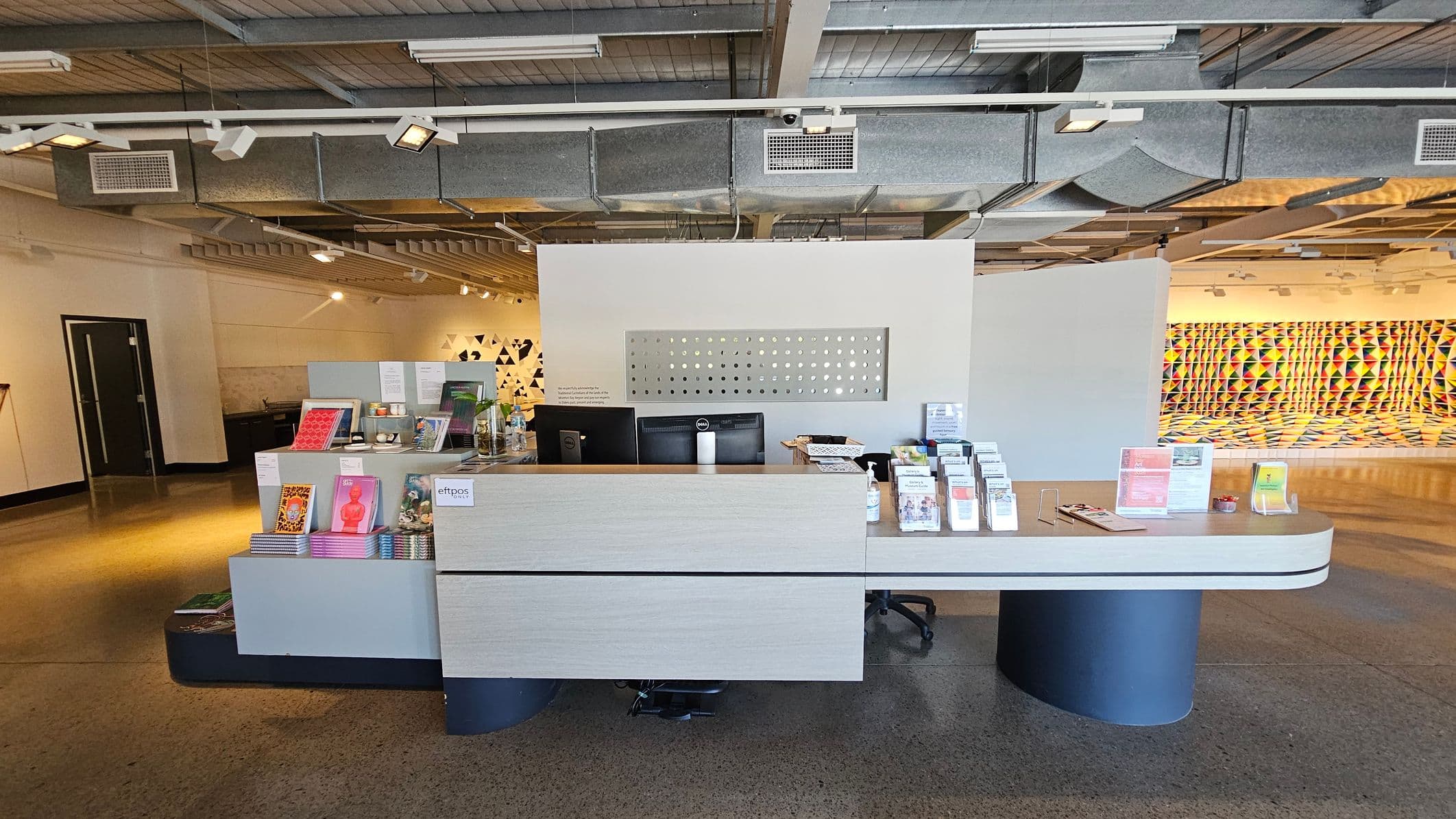 A spacious room with a polished concrete floor and an industrial-style ceiling featuring exposed beams and ductwork. On the right, a vibrant, geometric art installation with colourful triangular patterns covers the wall and extends onto the floor. In the middle, a white wall with a mounted screen and several stools. On the left, a desk with office chairs and a wall with a geometric design. The lighting is bright, highlighting the artwork.
