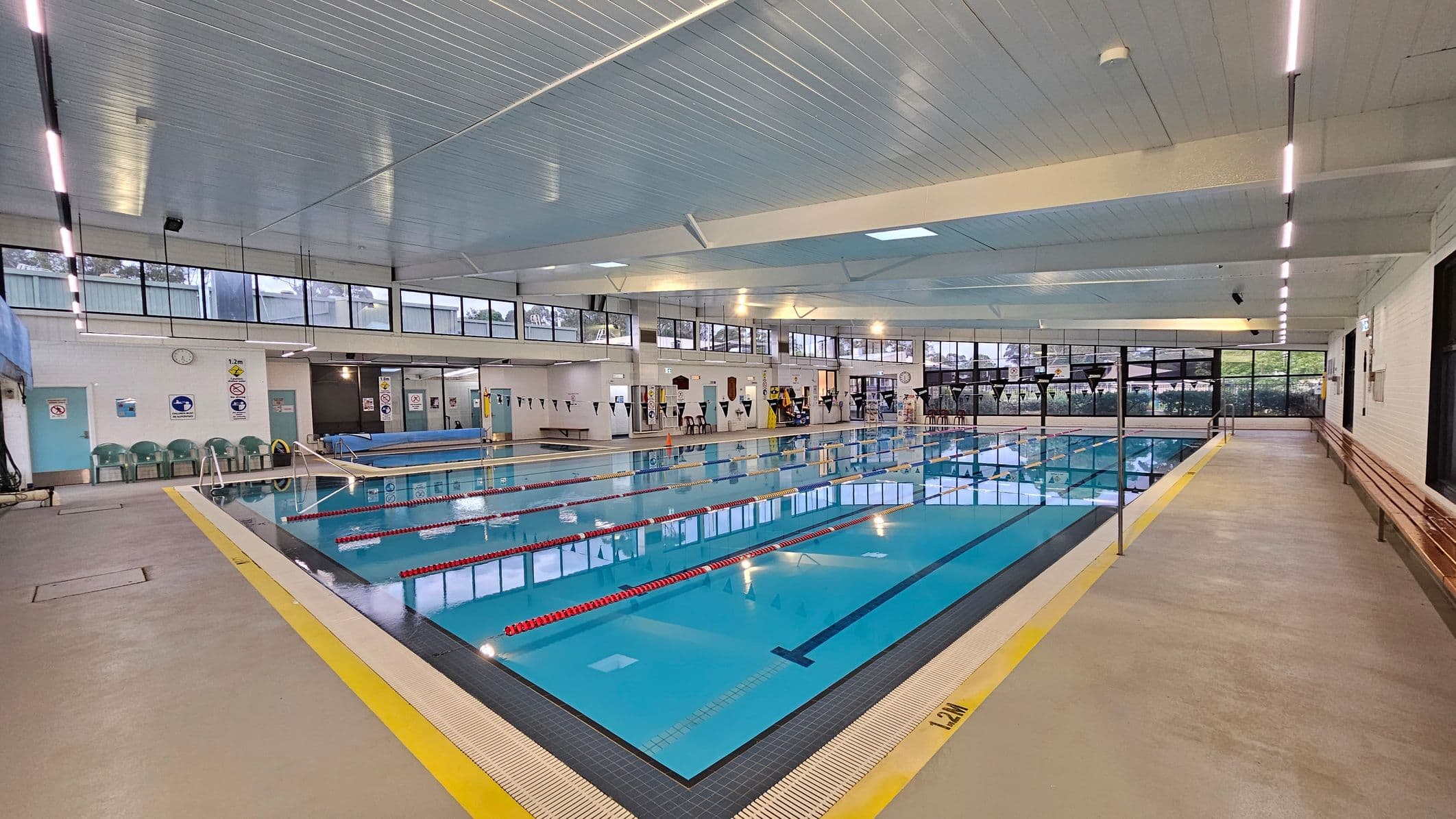An indoor swimming pool with clear blue water and red lane dividers. On the left, there are green plastic chairs against a white wall with signs and a clock. The right side features a long wooden bench along the wall. The ceiling is white with strip lighting, and large windows line the upper walls, allowing natural light in. The floor is a light grey with a yellow safety line around the pool's edge.
