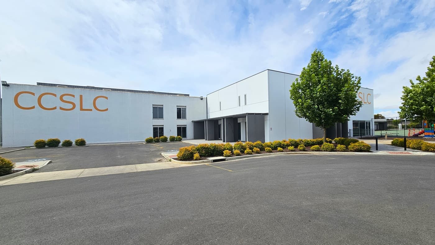 Copper Coast Sport and Leisure Centre is a large, white, and grey industrial building with a bold yellow-orange “CCSLC” sign on the left and front of the façade. The centre has glass entry doors and windows with white metal frames. Close to the left of the building are accessible car parking spaces, trees, and gardens with bushes maintained at a similar height. Right of the pavement leading to the entrance is a black, low, iron fence.
