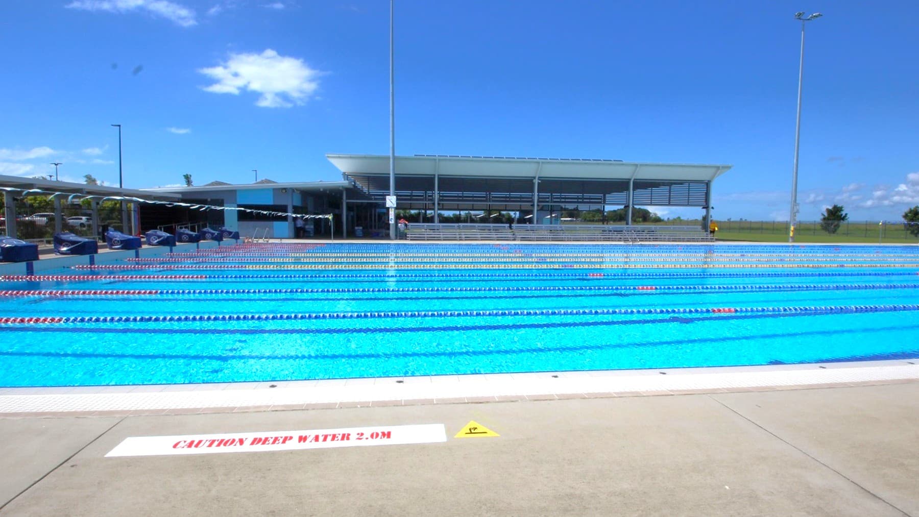 A 50m 10-lane pool features a ramp and stairs with handrails for entry. Diving boards with blue covers line both sides of the raised pool deck. Colourful floating rope lanes divide the pool, with white triangle banners hanging across. Tiered seating under a shade structure is positioned far left. Safety warnings adorn the concrete pool deck. Tall street lights illuminate the area.