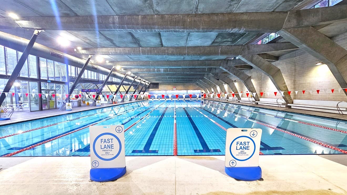A view from above of the 8 lane pool with small blue flags hanging across the pool. To the right are colourful murals along the wall and to the left people are making their way around the edge of the pool