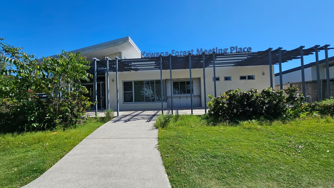 A modern building with a flat roof labelled "Kawana Forest Meeting Place" on the front. The structure has large windows and a pergola with vertical beams. In the foreground, there's a grassy area with a concrete path leading to the entrance. On the left, there are trees and shrubs, while the right side features more greenery and a wooden fence. The sky is clear and blue.
