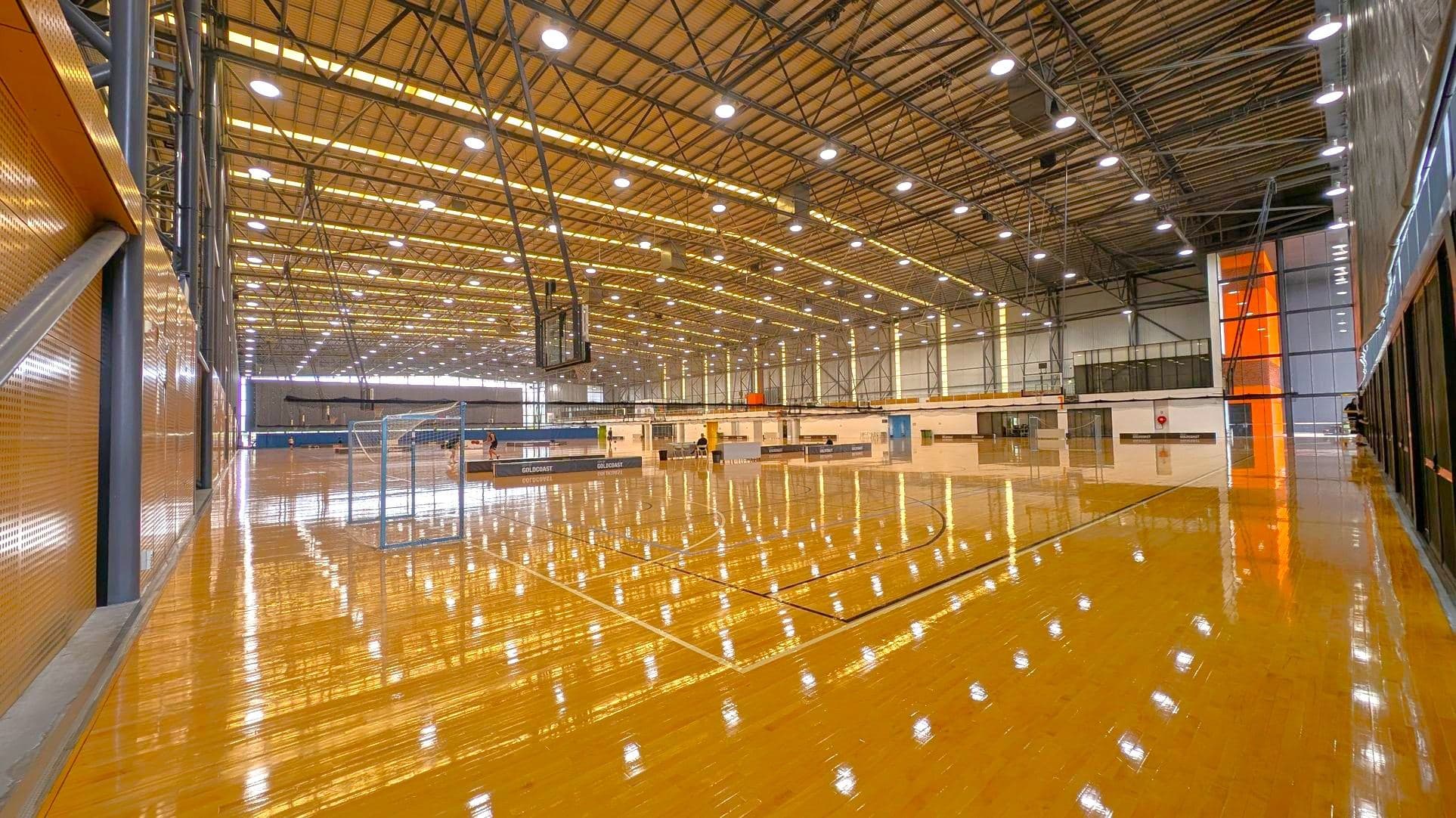 A large indoor basketball court with a polished wooden floor. Multiple basketball hoops are visible on the left and right sides. The ceiling is high with a metal framework and numerous lights. The background features large windows allowing natural light to enter. In the foreground, there are barriers with "CITY OF GOLD COAST" written on them. The walls are lined with light-coloured panels and some seating areas.