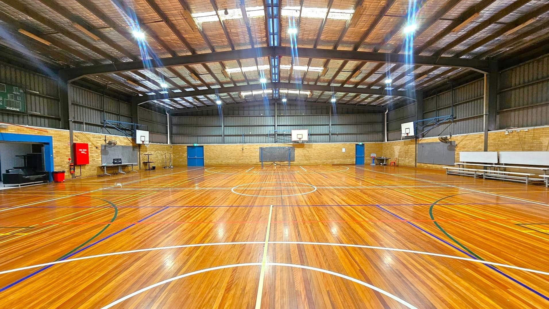 A spacious indoor basketball court with a polished wooden floor marked with various coloured lines. Two basketball hoops are mounted on the brick walls at either end. The ceiling is high with exposed beams and bright overhead lights. On the left, there is a scoreboard and a red fire hose reel. On the right, there are benches and a fan mounted on the wall. Blue doors are visible on both sides of the court.