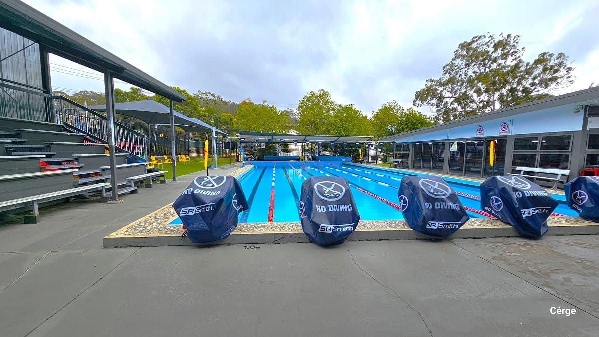 25m with 6 lanes outdoor aquatic pool at Mudgeeraba Aquatic Centre. The pool lanes are separated by red, white, and blue floating rope lanes. Two strings of triangular blue flags are tied around the opposite poles with a yellow flotation device. Seen at the back of this image are diving platforms covered with a blue cloth at the start of each pool lane, and to the right is multi-tiered seating. Yellow, blue, black, and grey plastic chairs are available all throughout the area.