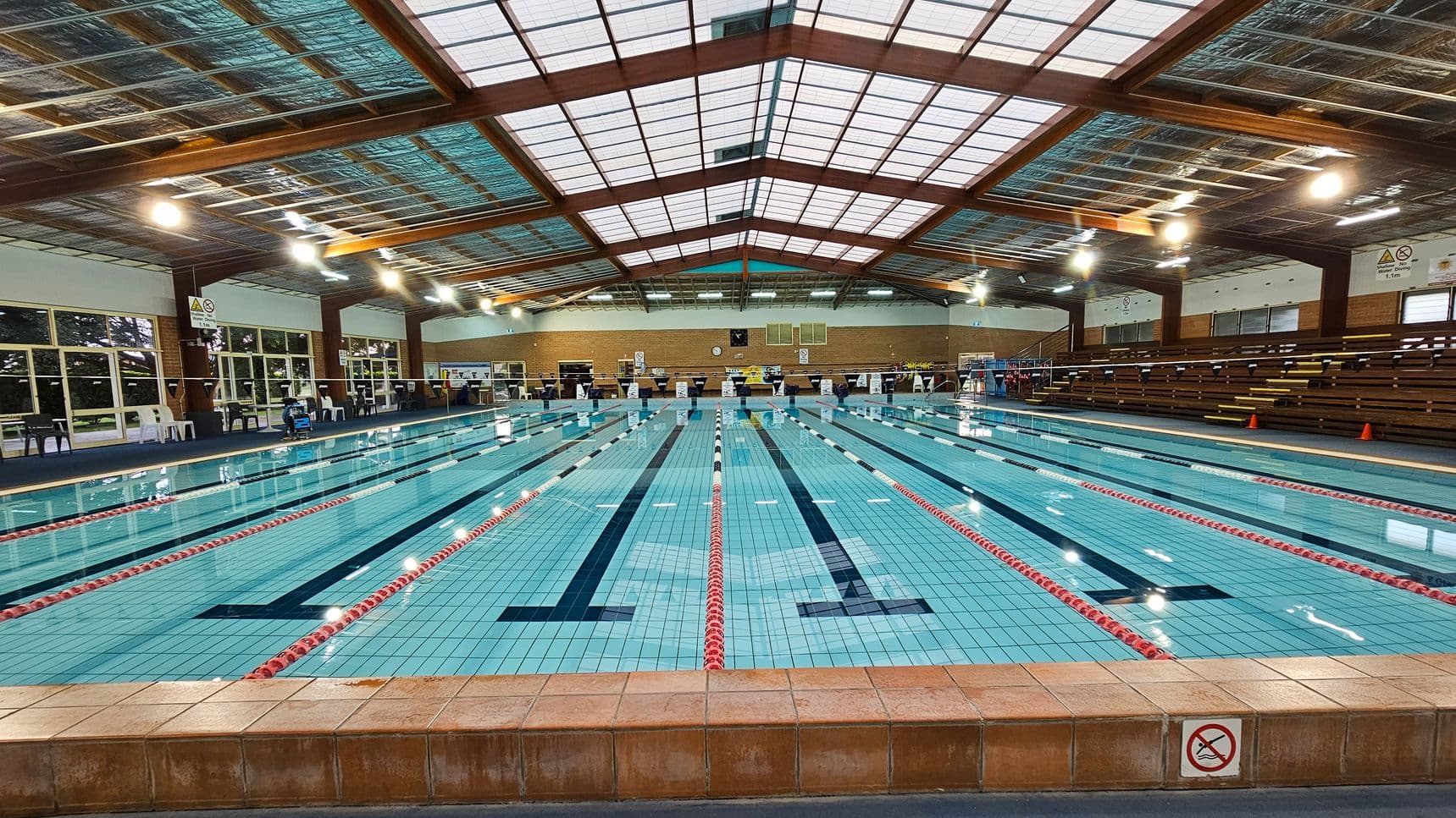 An indoor swimming pool with multiple lanes, separated by red and white lane dividers. The pool is surrounded by a tiled deck. On the right, there are wooden bleachers for spectators. The left side features large windows and several white plastic chairs. The ceiling is made of metal and wood beams, with bright overhead lights illuminating the area. The back wall has a clock and various signs.