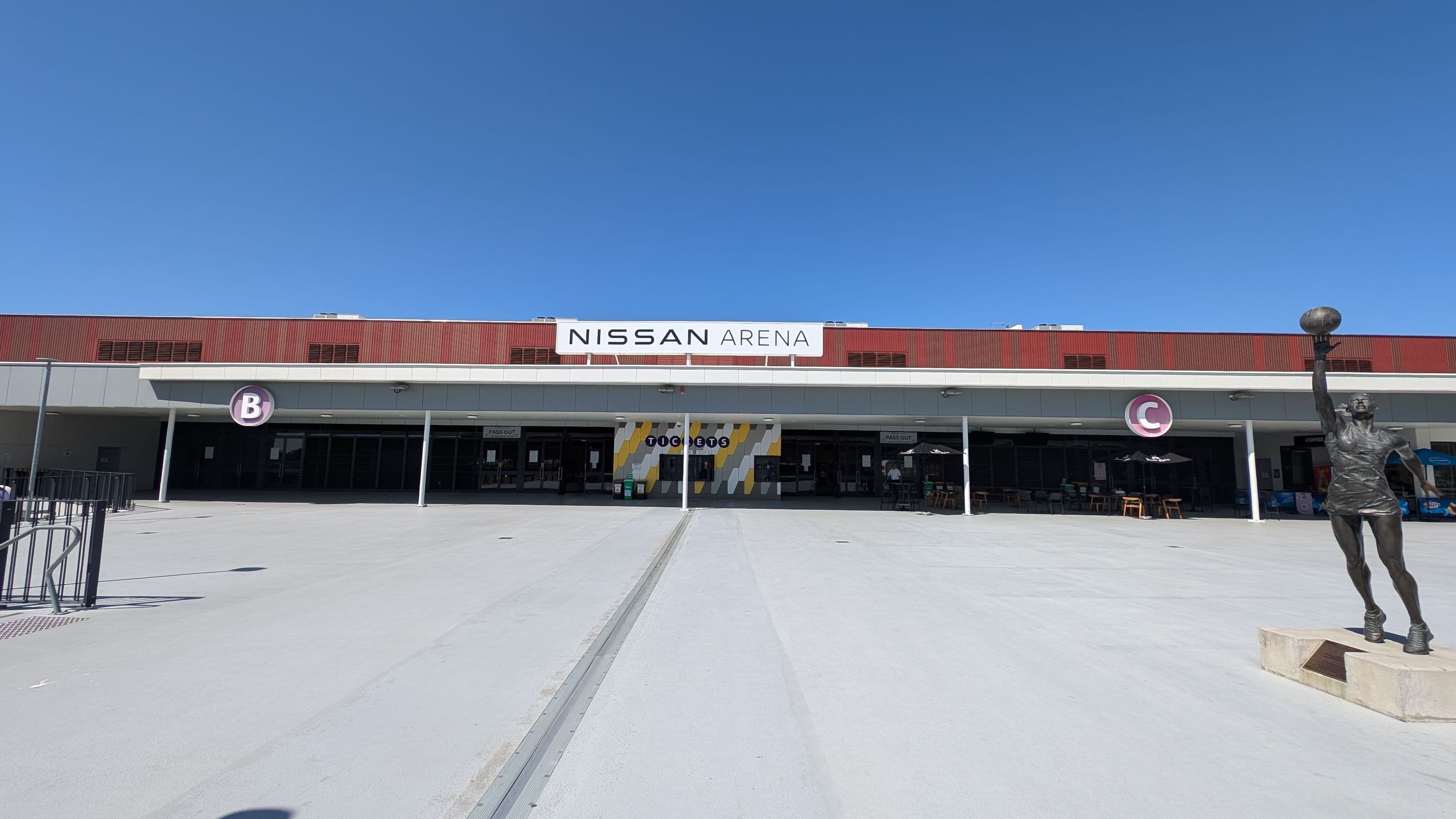 A wide view of the Nissan Arena entrance. The building has a red and white facade with a large sign reading "Nissan Arena" at the top centre. Below, there are entrances marked with letters "B" and "C". A statue of a person holding a ball is on the right side in the foreground. The ground is paved, and there are some outdoor seating areas with umbrellas near the entrances.