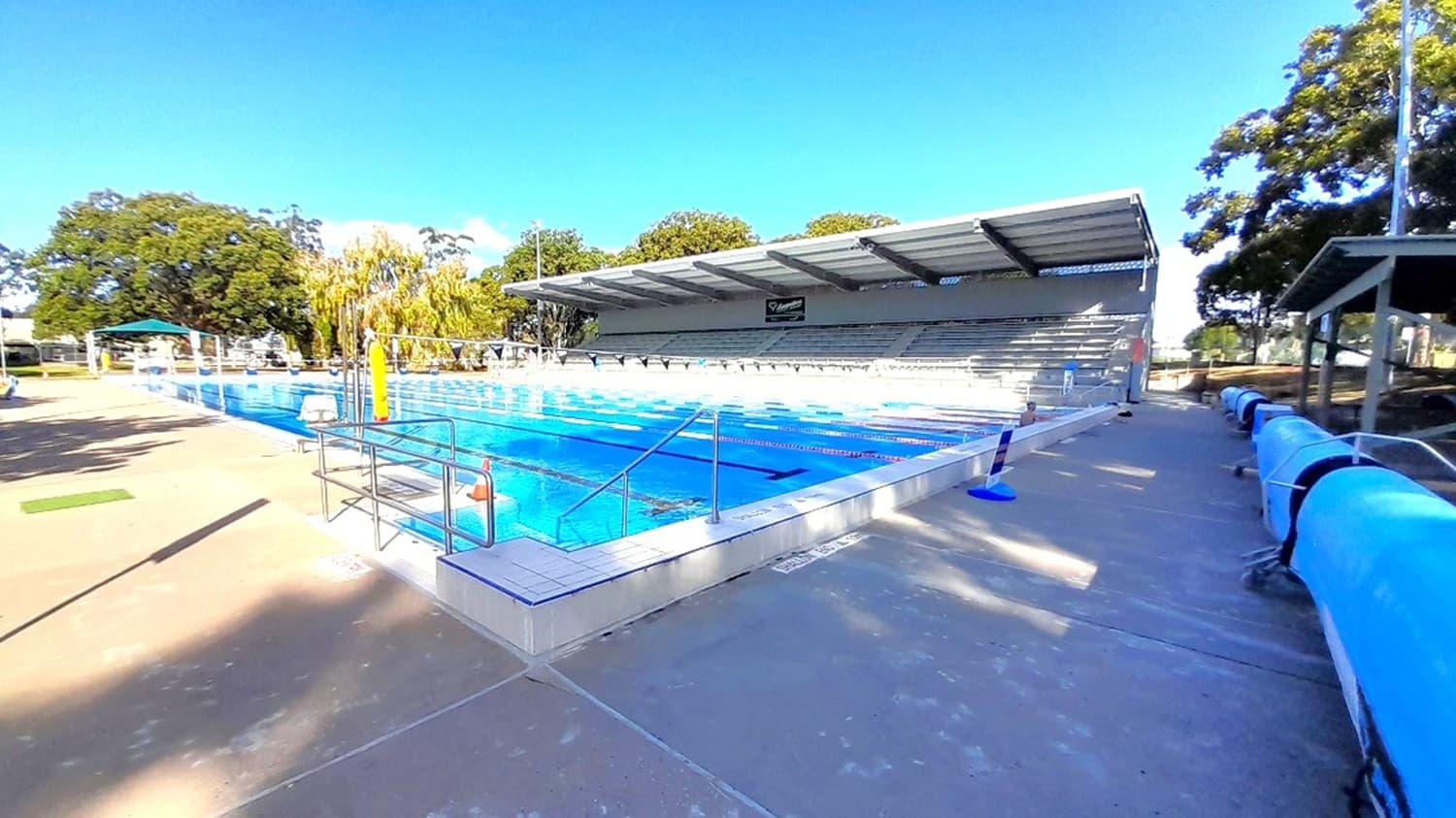A view over looking the outdoor pool, with a row of small blue flags hanging above the pool. In the background is the grandstand, with tiered seating.