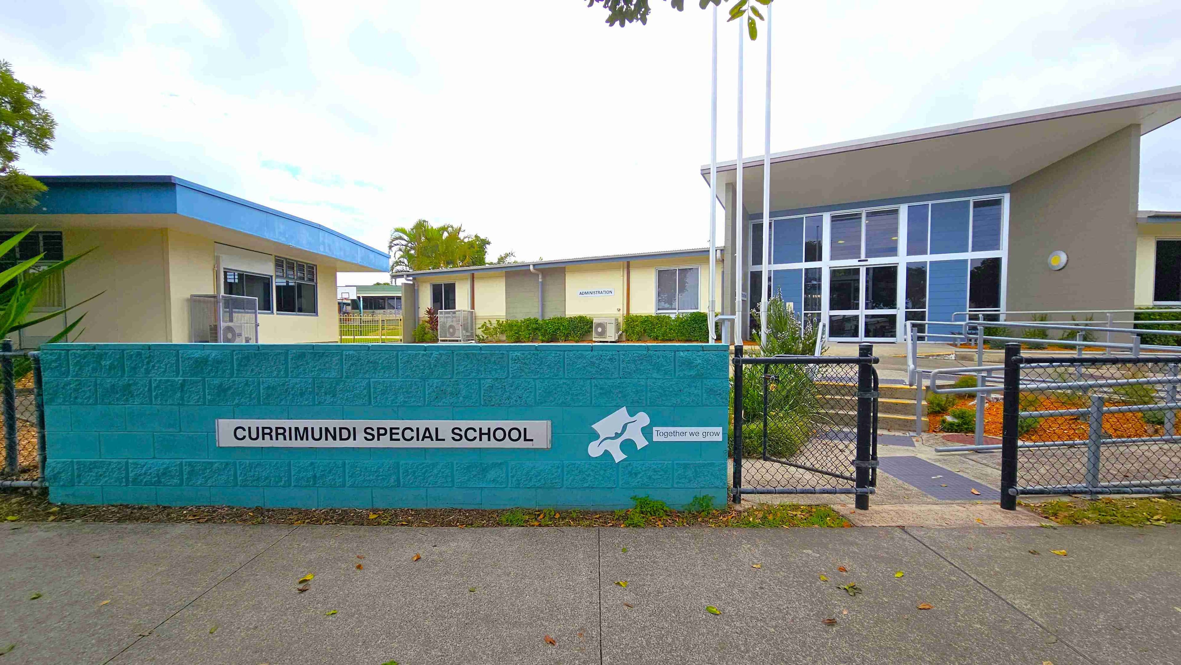 A school building with a modern design is on the left, featuring large windows and a sign reading "Administration" In front, a teal brick wall displays the name "Currimundi Special School" Three flagpoles stand nearby. On the right, a paved footpath runs alongside a fence, bordered by trees and a mulched area. Cars are parked in a lot further to the right