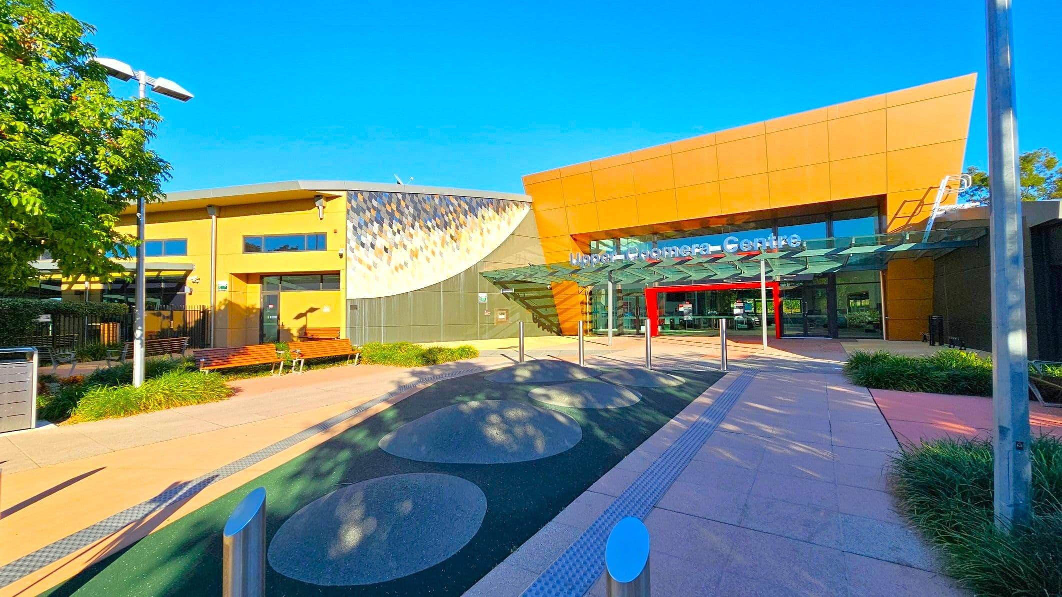 A modern building with a glass entrance is on the right, featuring a sign above the door. The structure has a geometric design with a mix of tan and grey panels. On the left, there is a smaller building with a mosaic wall and several windows. Benches and greenery line the walkway, which has circular patterns on the ground. A tall lamp post and a tree are visible on the left, and the area is well-lit by sunlight.