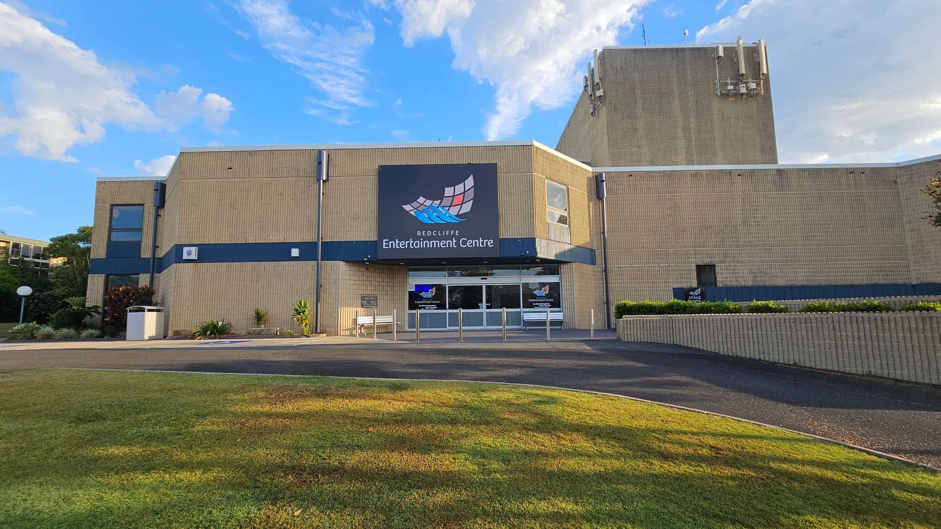 A beige brick building with a large sign reading "Redcliffe Entertainment Centre" above the entrance. The entrance has glass doors and is flanked by small plants and a bench on the left. The building has a blue horizontal stripe and several small windows. In the foreground, there is a grassy area leading to a paved path. The sky is visible above the building.