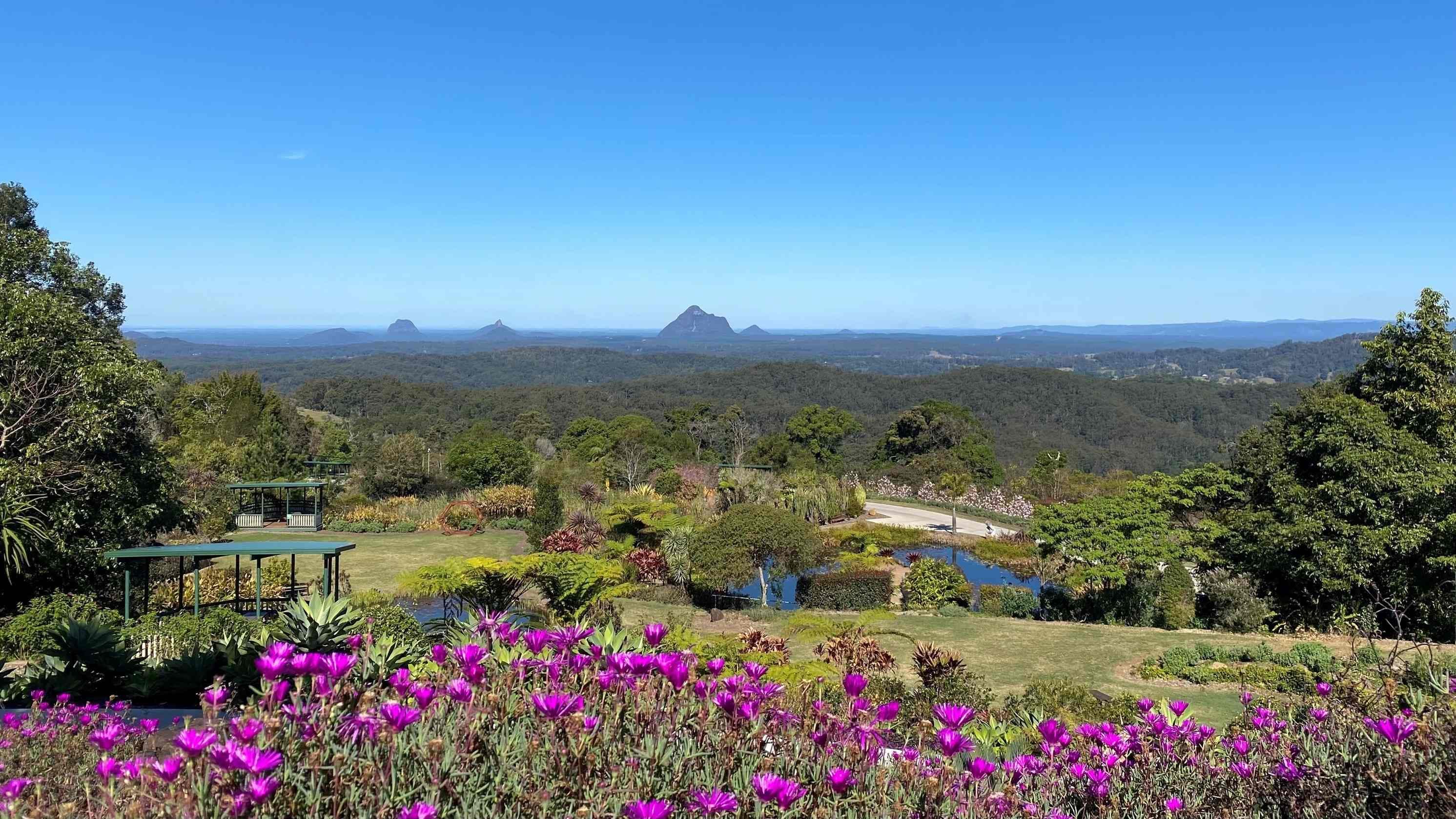 In the foreground, vibrant purple flowers are in full bloom. To the left, there is a small gazebo, surrounded by lush greenery. On the right, dense trees and shrubs extend towards the horizon. In the middle, rolling hills and valleys stretch out, leading to distinct mountain peaks in the distance. The background features a vast expanse of forested landscape.