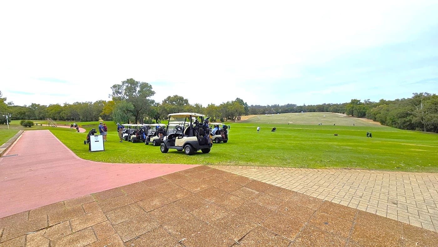 To the right, a pink concrete path descends to the greens, separated by a concrete border that extends across the putting green area. On the right, a sloping hill is covered in green grass. Further ahead, the concrete path continues to the other section of the golf area. The terrain varies in depth and elevation and is surrounded by trees. Golf players are visible in the distance.