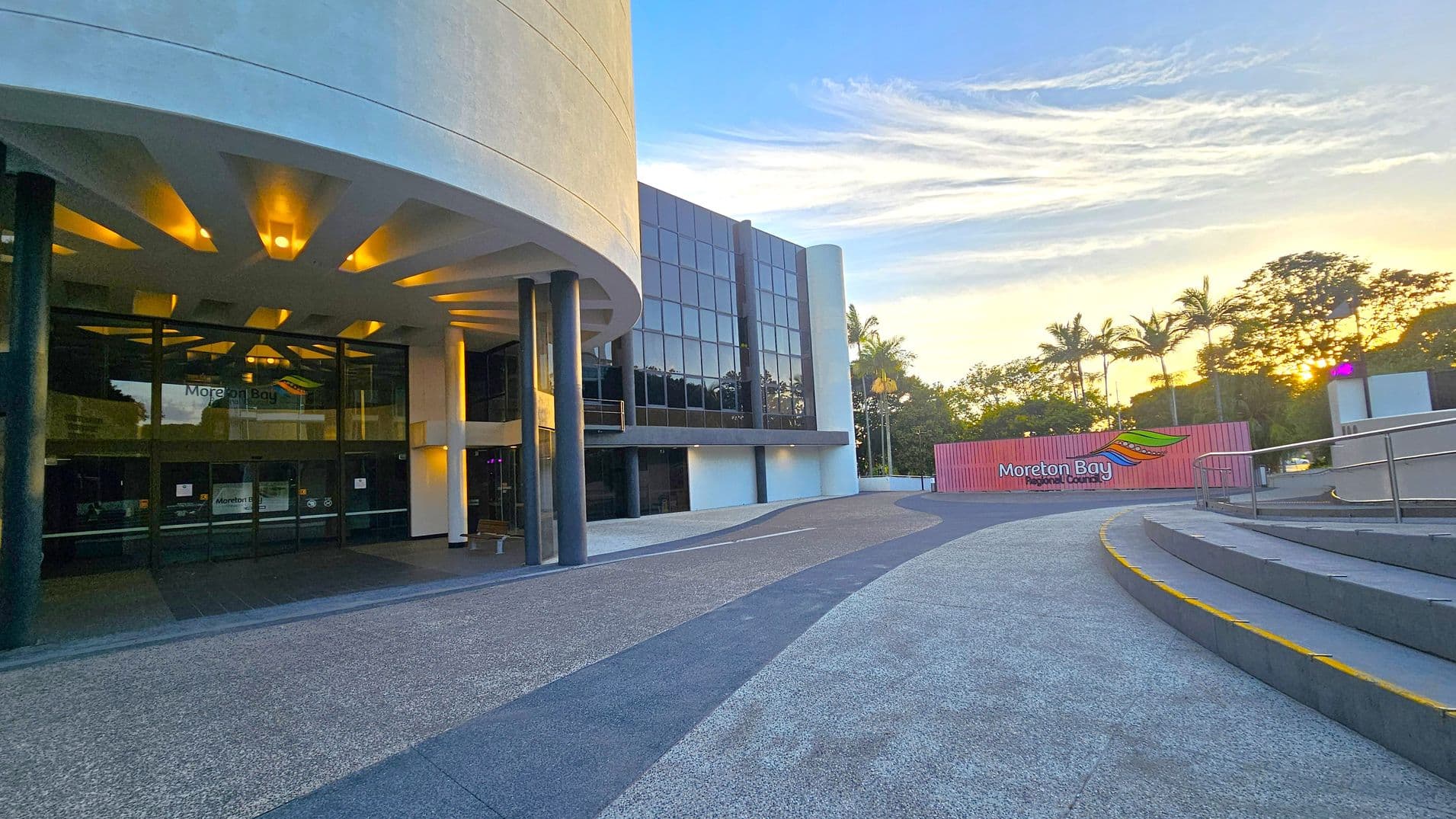 On the left, a modern building with a curved facade and large glass entrance is illuminated by warm lights. The building has tall, dark columns supporting a white overhang. On the right, a sign reads "Moreton Bay Regional Council" with a colourful logo. The foreground features a textured pathway and steps with yellow edges. In the background, palm trees and other foliage are silhouetted against a sunset sky.