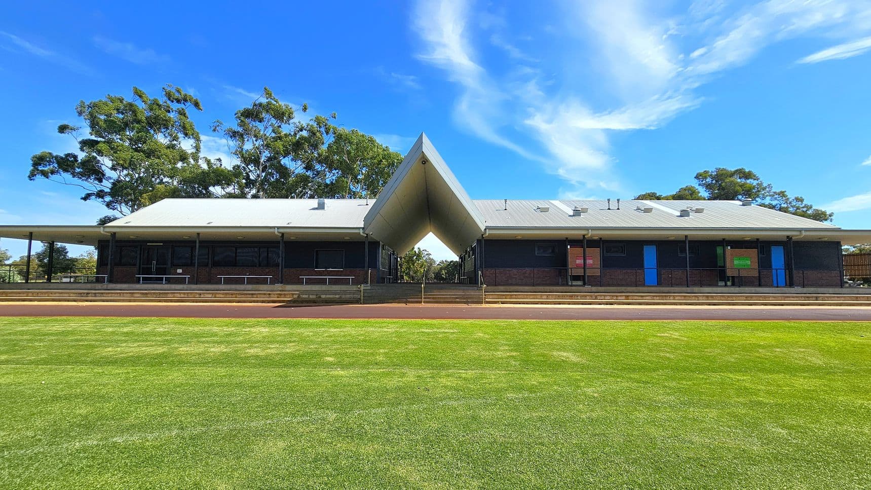 A building with a distinctive V-shaped roof structure in the centre, flanked by two sections with grey roofs. The building has dark walls with blue and green doors. In the foreground, there is a well-maintained grassy area. Trees are visible in the background on both sides.