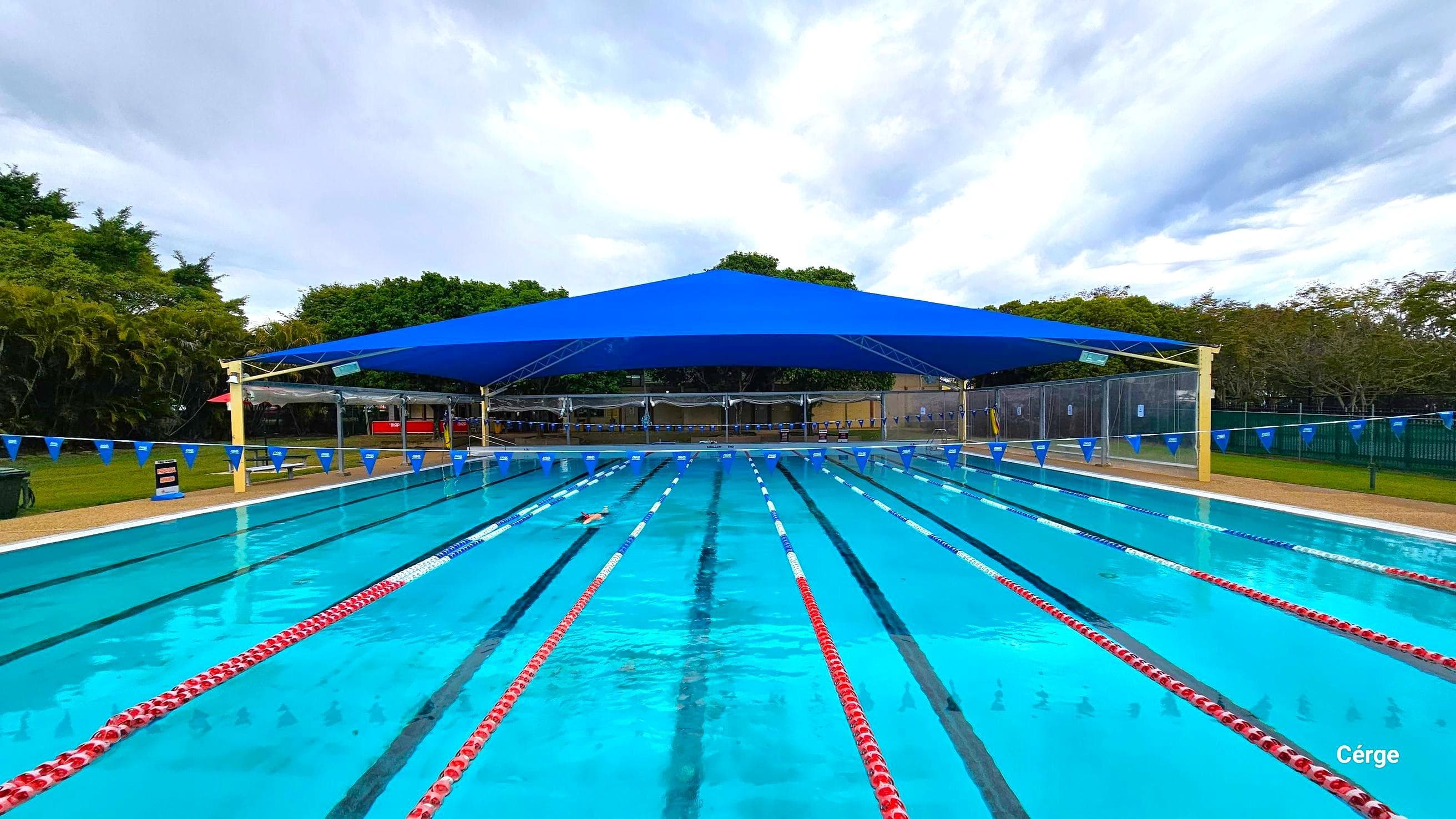 25m, nine-lane outdoor pool at Murrumba Downs Swimming Pool. The starting pool section has an elevated grey tile floor with nine white diving blocks that have orange traffic cones on top. The pool deck is brown with a rough surface to avoid slipping. The other end is partially covered by a wide blue sunshade supported by yellow metal frames. A few benches with tables are available around the pool, and strings of triangular blue flags are wrapped around steel posts on opposite pool sides.
