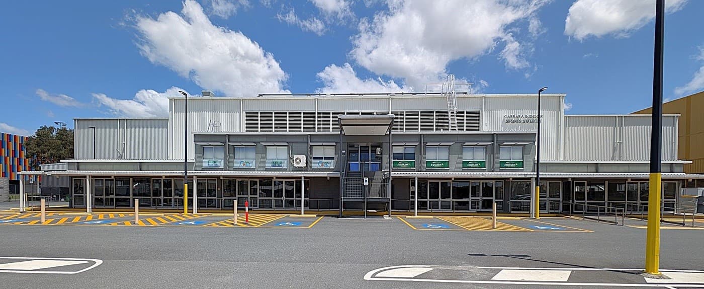 A large, modern building with a grey facade and multiple glass doors at the front. The upper section has several windows with green awnings. The foreground features a spacious car park with marked parking spaces, including accessible spots with blue and yellow markings. A colourful structure is partially visible on the left side, and there are several tall lamp posts around the area. The ground is paved with asphalt.