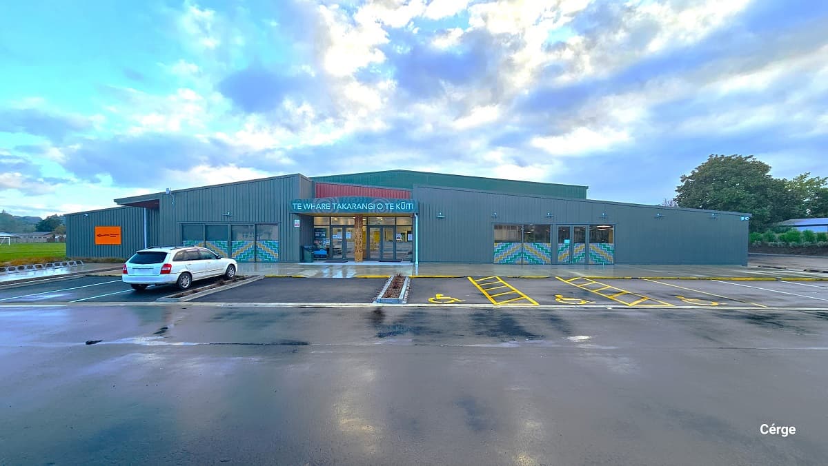 Gallagher Recreation Centre is a low-set with a grey corrugated structure, glass windows, and orange, brown, and green features. At the upper part of the main entrance is an illuminated text. On the left upper side wall is a ‘no smoking sign.’ There is a large space in front of the facility, and several residential houses can be seen in the surrounding area.