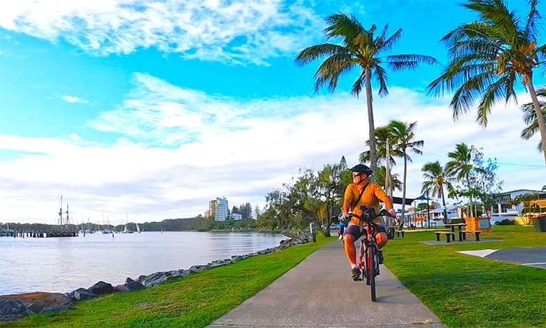 On the left, a dark blue electric bike with a helmet hanging on the handlebars. On the right, a green electric bike with a similar design. In the middle, both bikes are standing on their kickstands, facing slightly towards each other. The background features a grassy park area with tall trees and some buildings in the distance.